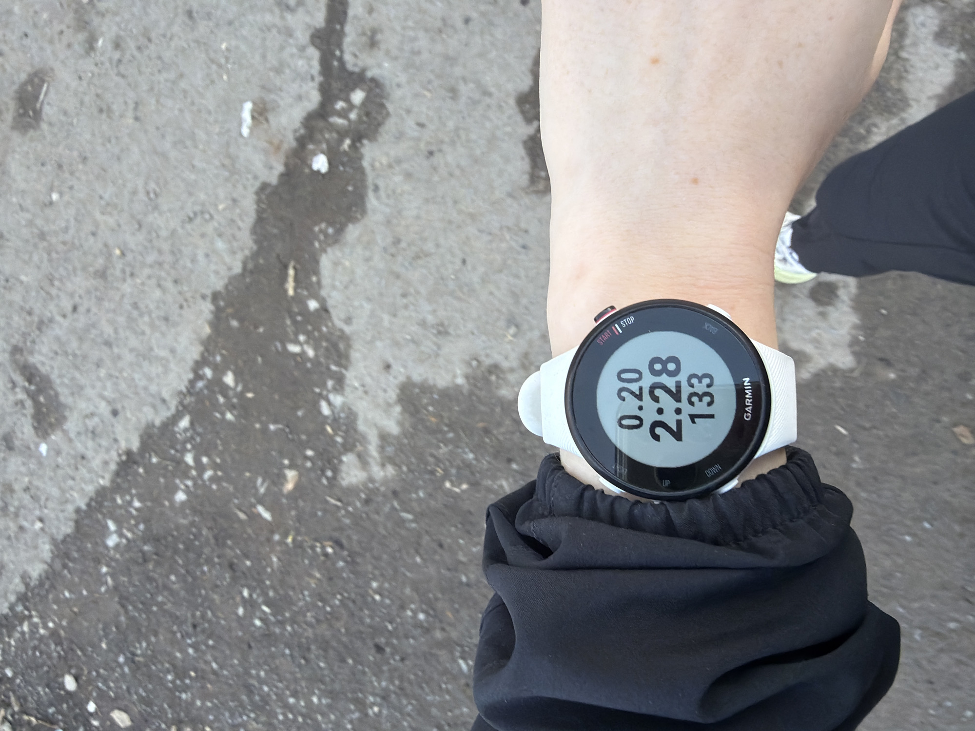 A first-person perspective photograph shows a person's wrist with a white Garmin smartwatch displaying 0.20, 2:28, and 133. The person is wearing a black long-sleeved garment. Part of a running shoe with white and lime-green details is visible. The background consists of a concrete surface, possibly a sidewalk or road, with a dark, wet-looking area on the left. There is no information to indicate that this photo was taken in Berezniki, Russia.