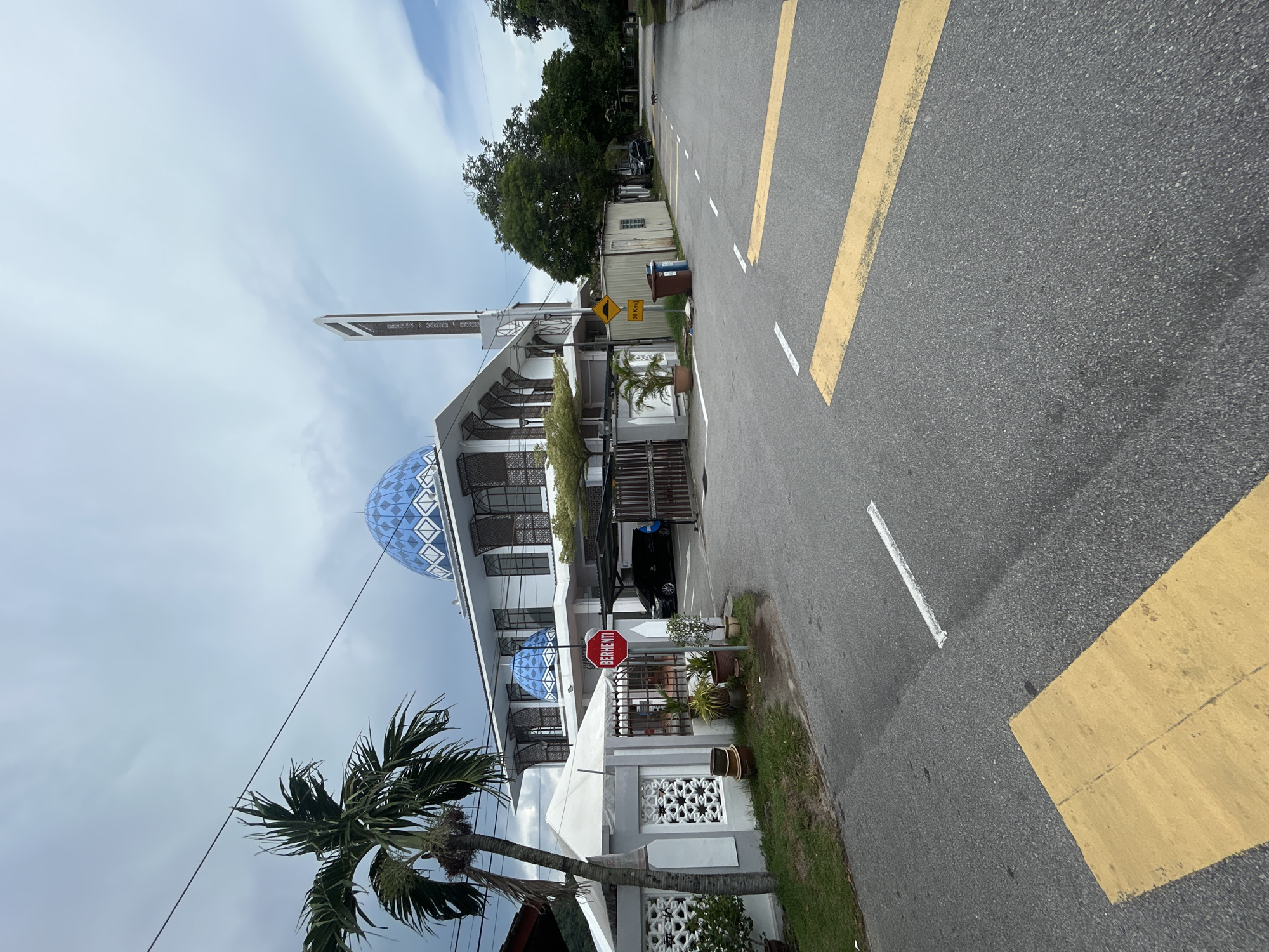 This is an exterior, eye-level, medium shot of a mosque in George Town, Malaysia, on a cloudy day. The mosque features a prominent blue dome and a tall, slender minaret. The building is white with ornate windows and decorative elements. In the foreground, a paved road with yellow and white markings runs towards the mosque. A palm tree stands to the left of the frame, and a white canopy-like structure is also visible. A red stop sign with the word "BERHENTI" is posted near the entrance of the mosque, along with several potted plants. To the right of the mosque, a narrow street leads away from the camera, with trees lining its sides. Two trash bins are located near a small, metallic structure. A black car is parked under a carport to the right of the mosque entrance.