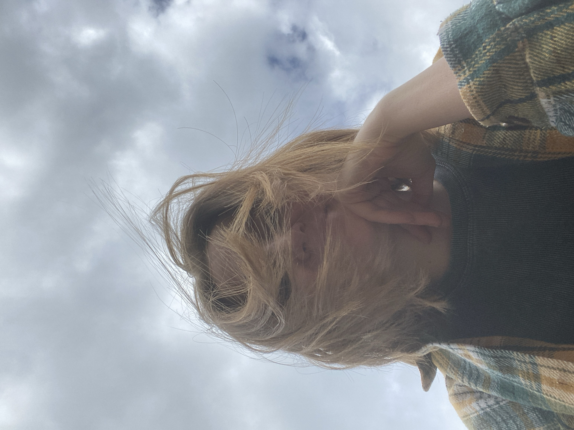 A close-up, low-angle selfie of a young person with blonde hair, partially obscured by windblown strands, leans their chin on their hand. They are wearing a dark grey ribbed shirt beneath an open plaid flannel jacket in green, yellow, and white. The background is a cloudy sky. The location is not identifiable from the image.