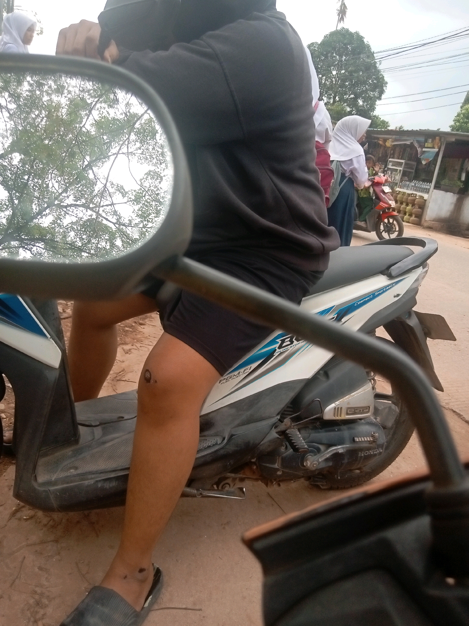 In Batam, Indonesia, the photograph captures a close-up view of a person sitting on a scooter, with a focus on their right leg and the scooter's body. The person is wearing dark shorts and black flip-flops. Their right knee displays a dark, circular mark, possibly a wound or birthmark. The scooter is white and blue, with the "HONDA" logo visible on the engine casing.
In the background, a red scooter is parked, with two individuals on it. One is wearing a white hijab and a school uniform, while the other is partially visible. To the right of the scooters, a shop with shelves stocked with bottles and other items is present. Trees and utility lines are visible in the distance, suggesting an outdoor, possibly semi-urban setting. The overall scene appears to be captured from the perspective of another person on a scooter, with a side mirror reflecting a blurry image of trees and sky.