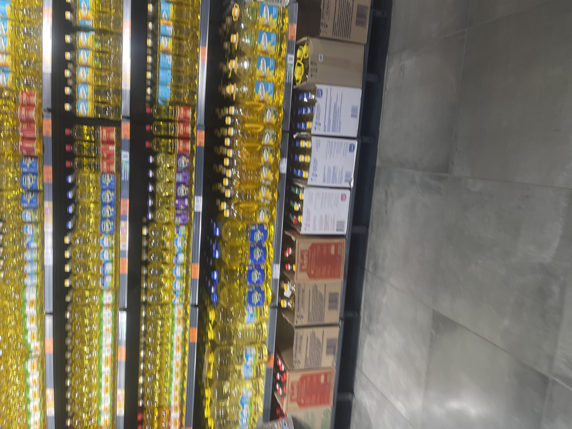 The image shows a retail store aisle with shelves stocked with cooking oil. The shelves are arranged horizontally, and there are multiple rows of bottles and boxes. The oil bottles are primarily clear with yellow contents, and some have labels with blue and white accents. There are also cardboard boxes containing bottles, some with red labels. The flooring is a polished concrete material. The lighting appears to be overhead fluorescent lights typical of a supermarket. No people are visible in the frame.