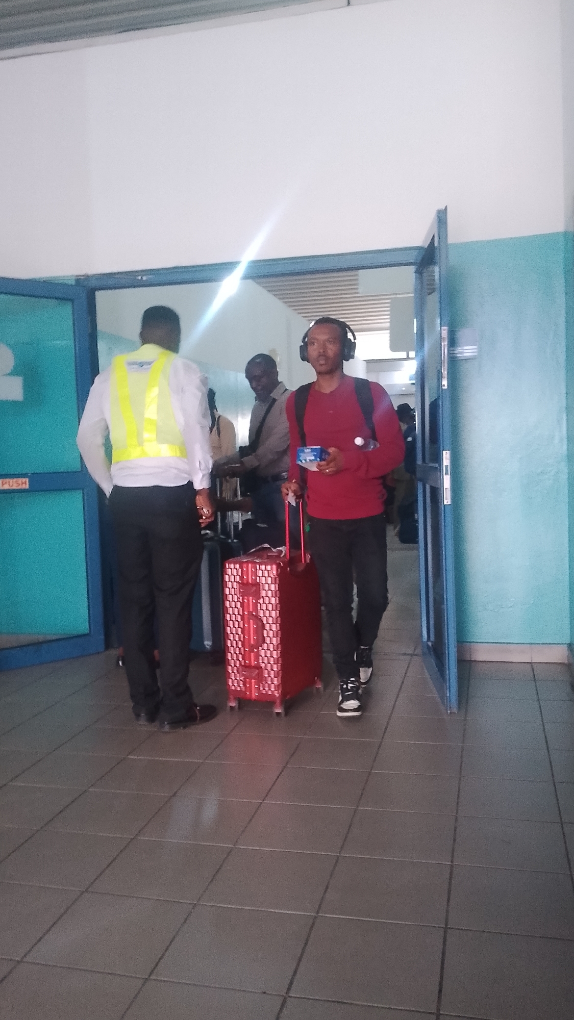 The image shows an indoor airport scene, presumably in Orerokpe, Nigeria. A man wearing a red shirt, black pants, and headphones is walking through a doorway pulling a red patterned suitcase. He is holding a small, rectangular item in his hand. Behind him, another man in a light-colored shirt and dark pants is standing, facing away from the camera. A security officer wearing a yellow vest is facing the same direction. Other people can be seen in the background near what looks like a baggage claim area. The floor is tiled. The doorway is blue and white with a "PUSH" sign visible on the left door. There's a bright glare of light towards the upper center of the image.