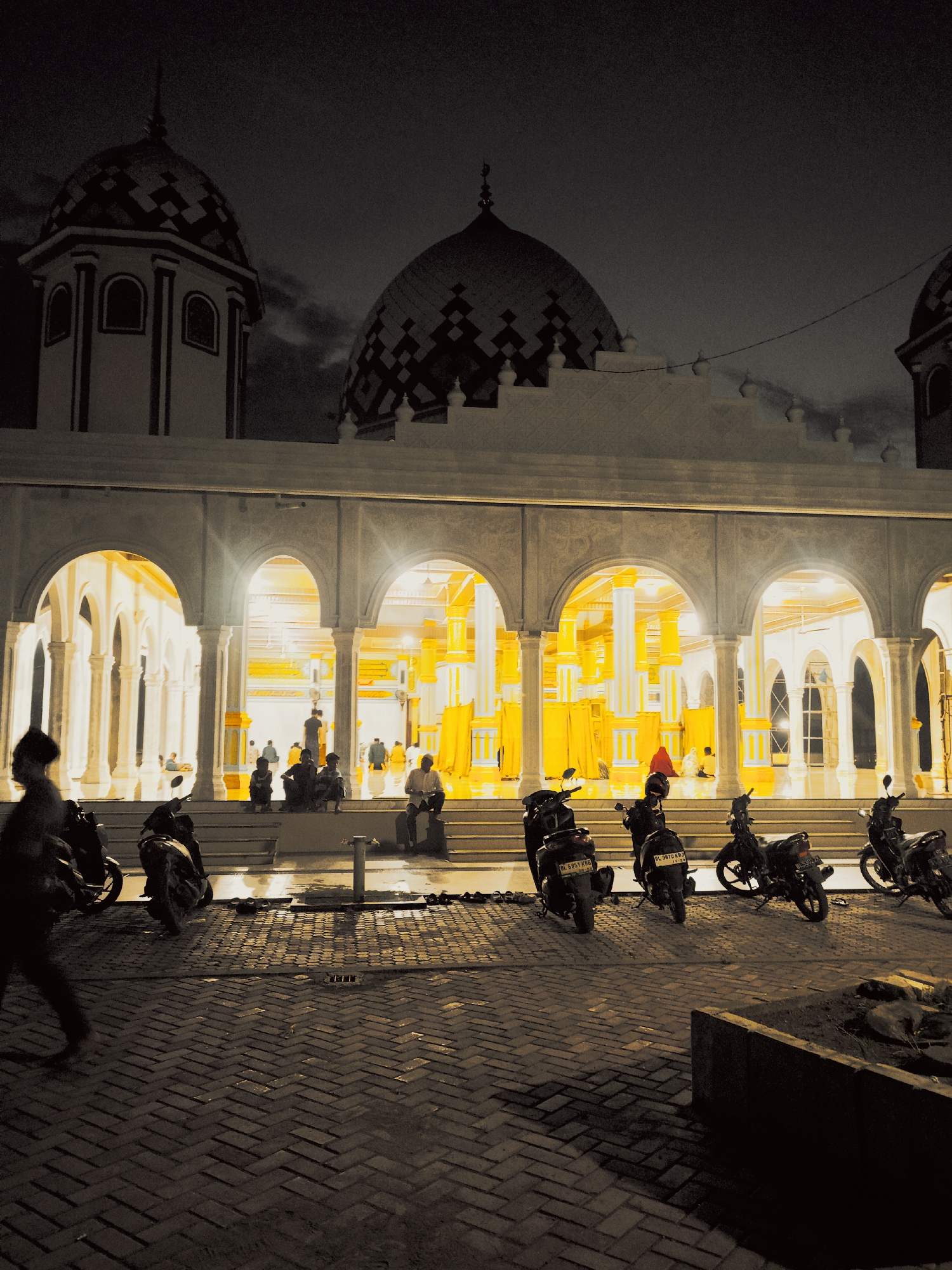 The image depicts the Baiturrahman Meunasahlhok Mosque at night in Meunasahlhok, Indonesia. The mosque features a white facade with multiple arches illuminated from within, revealing yellow columns and architectural details. Several people are seated on the steps leading into the mosque. A row of parked motorcycles occupies the foreground, with a person walking past on the left. The ground is paved with bricks. The mosque has domed roofs with patterned designs. The sky is dark, suggesting it is nighttime.