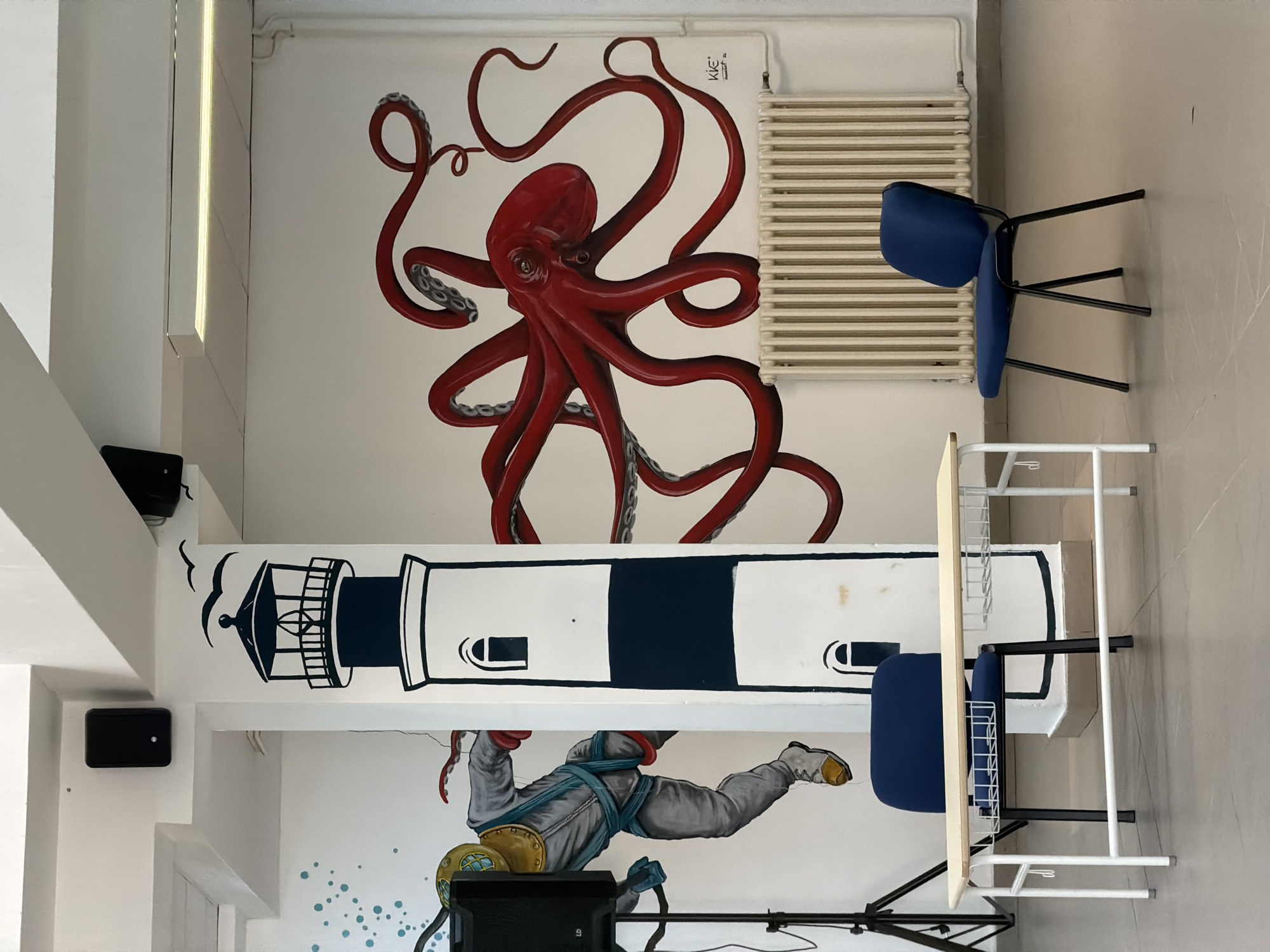 The image depicts a room with a large mural on the wall. The mural features a lighthouse, a diver in a vintage diving suit, and a giant red octopus. In the foreground, there is a white table with a light-colored wooden top and two blue chairs. One chair is placed at the table, and another is to the right of the table. A radiator is visible on the wall behind the right chair. The room appears to be an indoor space, possibly a classroom or a common area, with a light-colored floor. There are also speakers mounted on the ceiling. The overall scene is colorful and artistic, with a maritime theme suggested by the mural elements. The location of Budva, Montenegro is not directly discernible from the visual elements of the image.