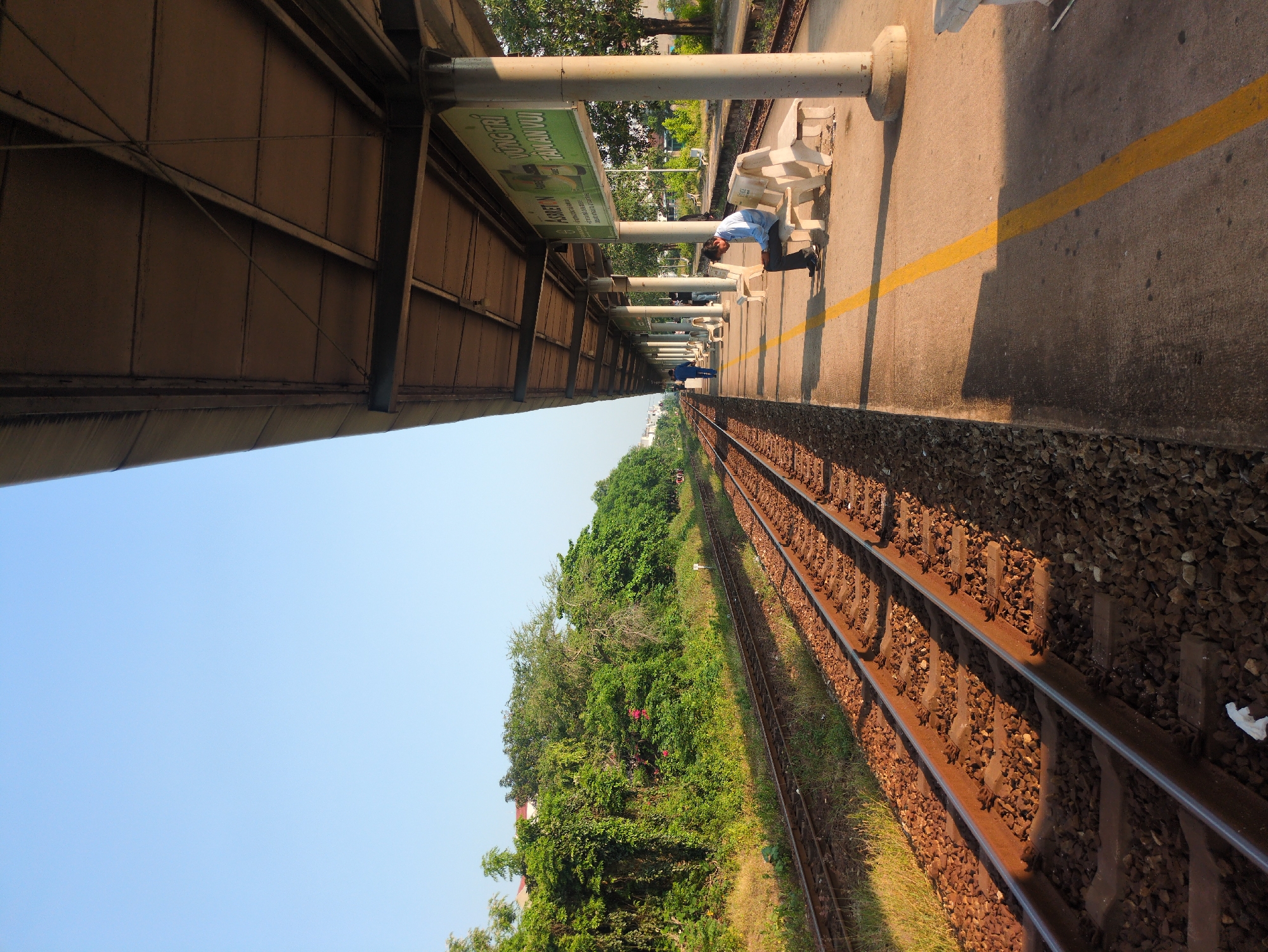 The image depicts a train station platform in Tuy Hòa, Vietnam. Parallel railway tracks run alongside the platform edge, covered in ballast stones. Lush green vegetation grows adjacent to the tracks. The platform is concrete and features a yellow safety line. White, modern-style benches are spaced along the platform, some occupied by people. A man in a blue shirt is bent over, interacting with something on the platform near a pillar. Overhead, the platform is covered by a metal canopy structure supported by concrete pillars. A green advertisement sign is mounted on one of the pillars. The sky is clear and blue.