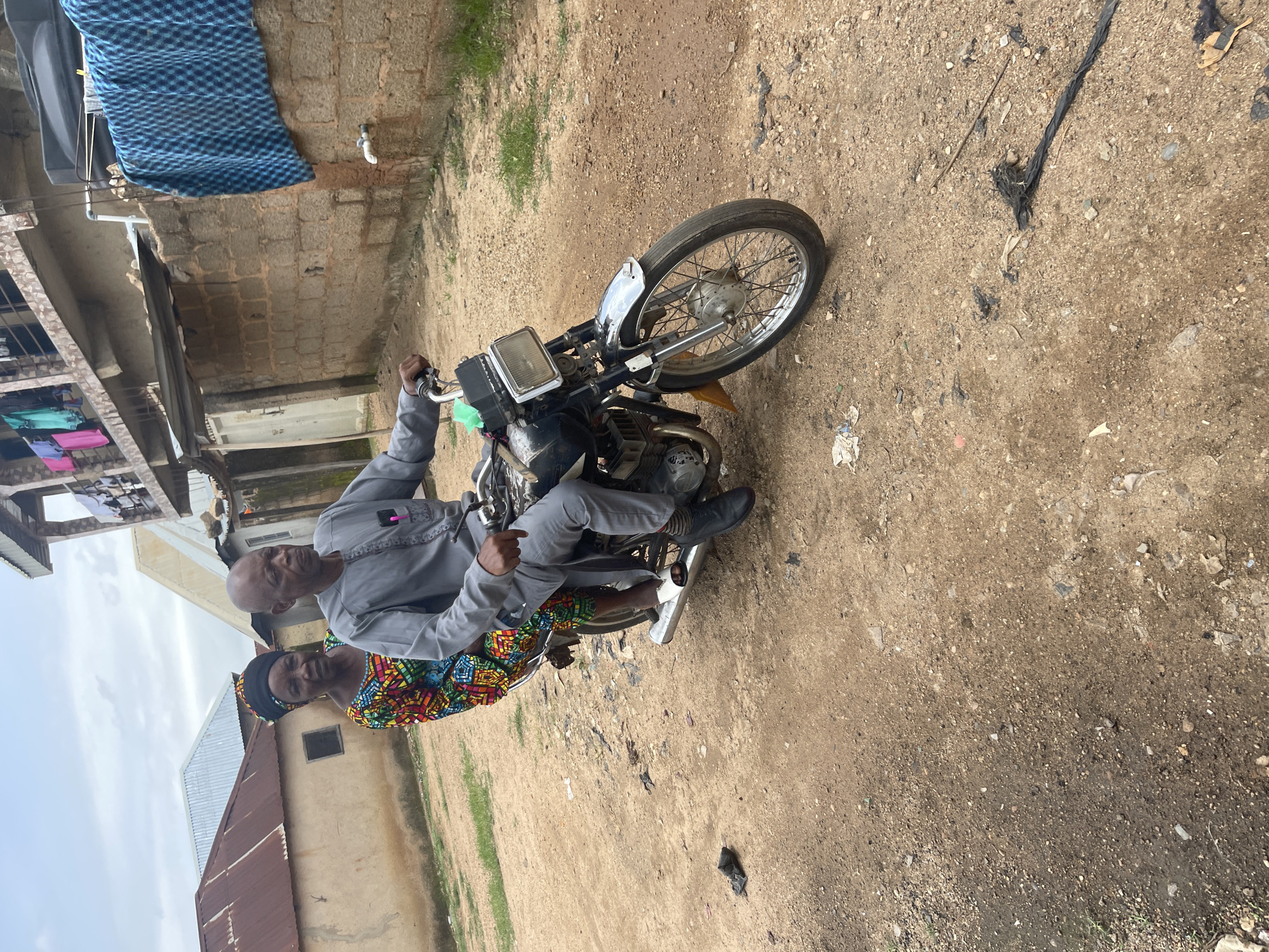 A man and a woman are riding on a motorcycle in Rafin Par, Nigeria. The man is driving the motorcycle, and the woman is sitting behind him. The man is wearing a gray outfit and the woman is wearing a colorful dress and a headscarf. The motorcycle is black and has a silver exhaust pipe. The ground is dirt and there are some buildings in the background. The sky is cloudy.