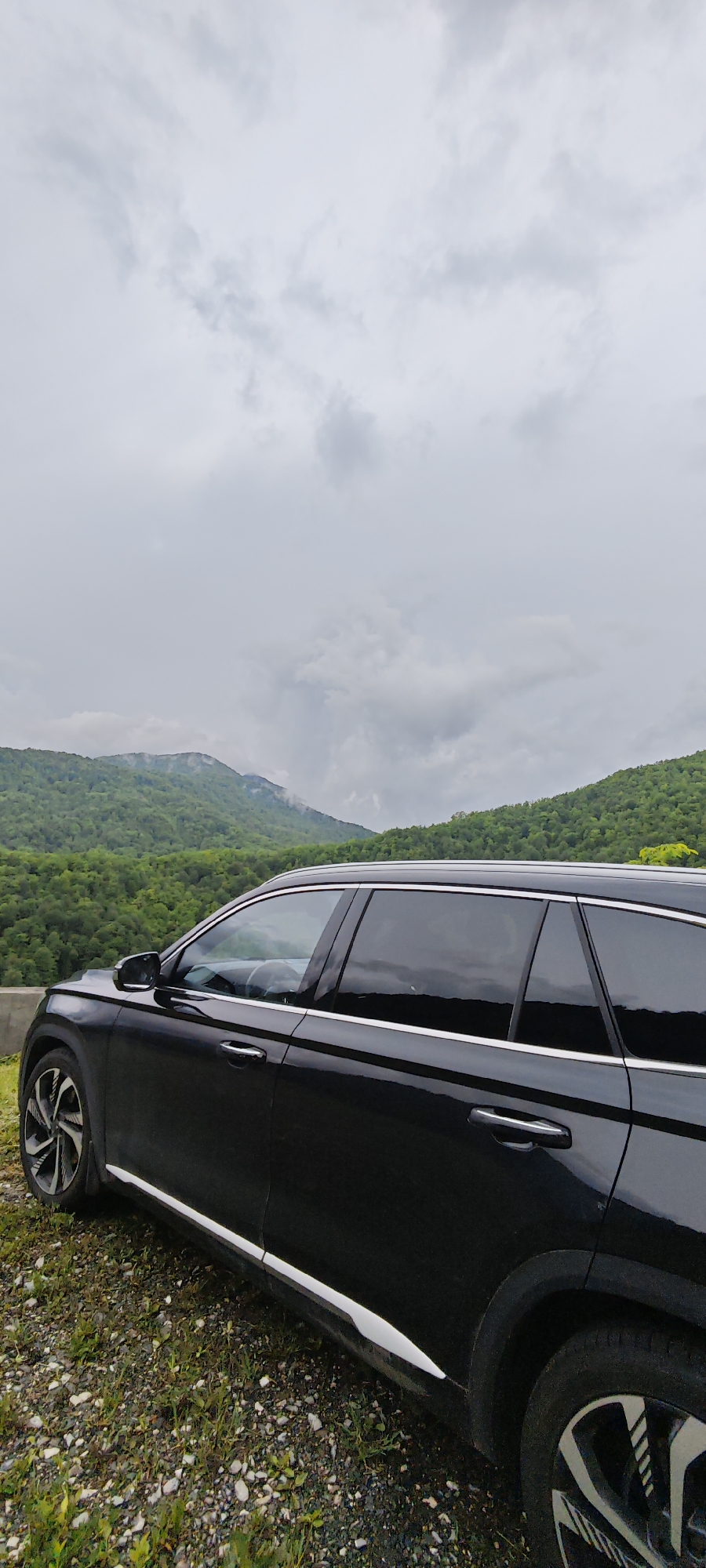 A black station wagon is parked on a ledge overlooking a green, mountainous landscape under a cloudy sky. The car is angled towards the viewer, with the driver's side visible. The landscape consists of trees covering the mountains, with some distant peaks partially obscured by clouds. The ledge where the car is parked appears to be covered in gravel and sparse vegetation. The location is Estosadok, Russia, as confirmed by the photo context.