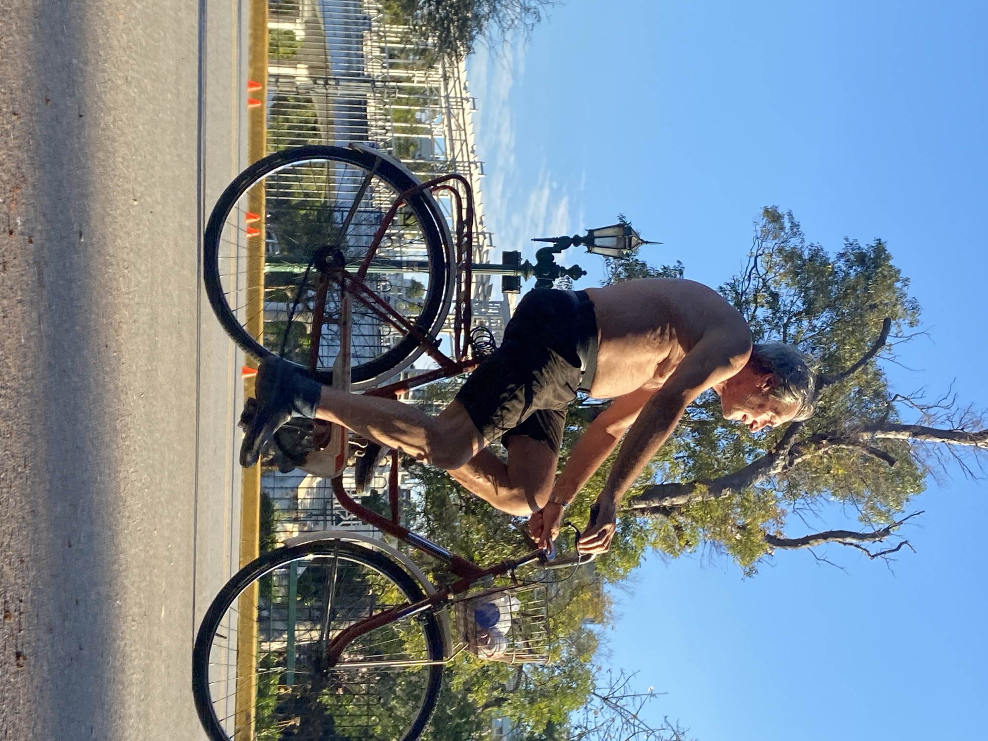An adult male is riding a bicycle on a city street. The man is fair-skinned, with grey hair, wearing black shorts and black shoes. He is shirtless, showing his torso. The bicycle is old, with a basket on the front and a carrier rack over the back wheel. A lamppost is visible behind the bicycle. The background includes a fence, trees, and a blue sky. Orange traffic cones line the side of the road. The image location is Buenos Aires, Argentina.