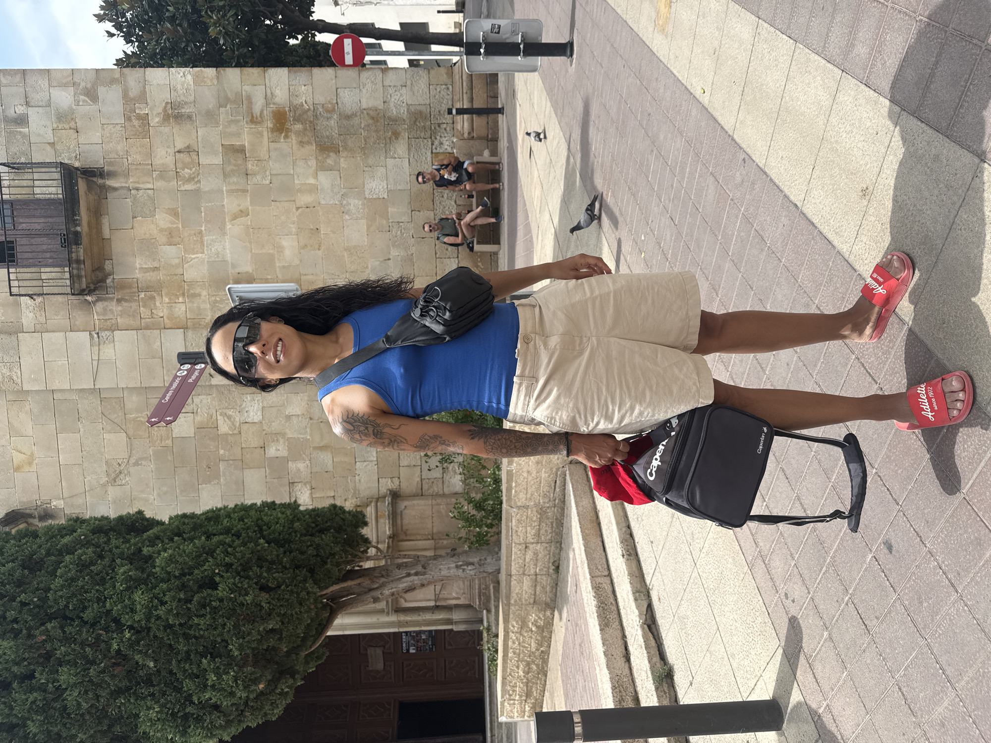 A woman stands on a paved sidewalk in front of a large, stone building with arched doorways and windows. She is wearing a blue tank top, tan shorts, and red slide sandals. A black crossbody bag is slung over her shoulder, and she is holding a black backpack with a red item peeking out from the top. She has sunglasses on and her hair is dark and curly. In the background, two men sit on a bench, and pigeons are visible on the ground. A street sign indicates directions to "Centre Historic" and "Platges" (Historic Center and Beaches), consistent with Tossa de Mar, Spain, a coastal town known for its well-preserved medieval architecture. The lighting suggests it is daytime, likely a warm day given the clothing choices.