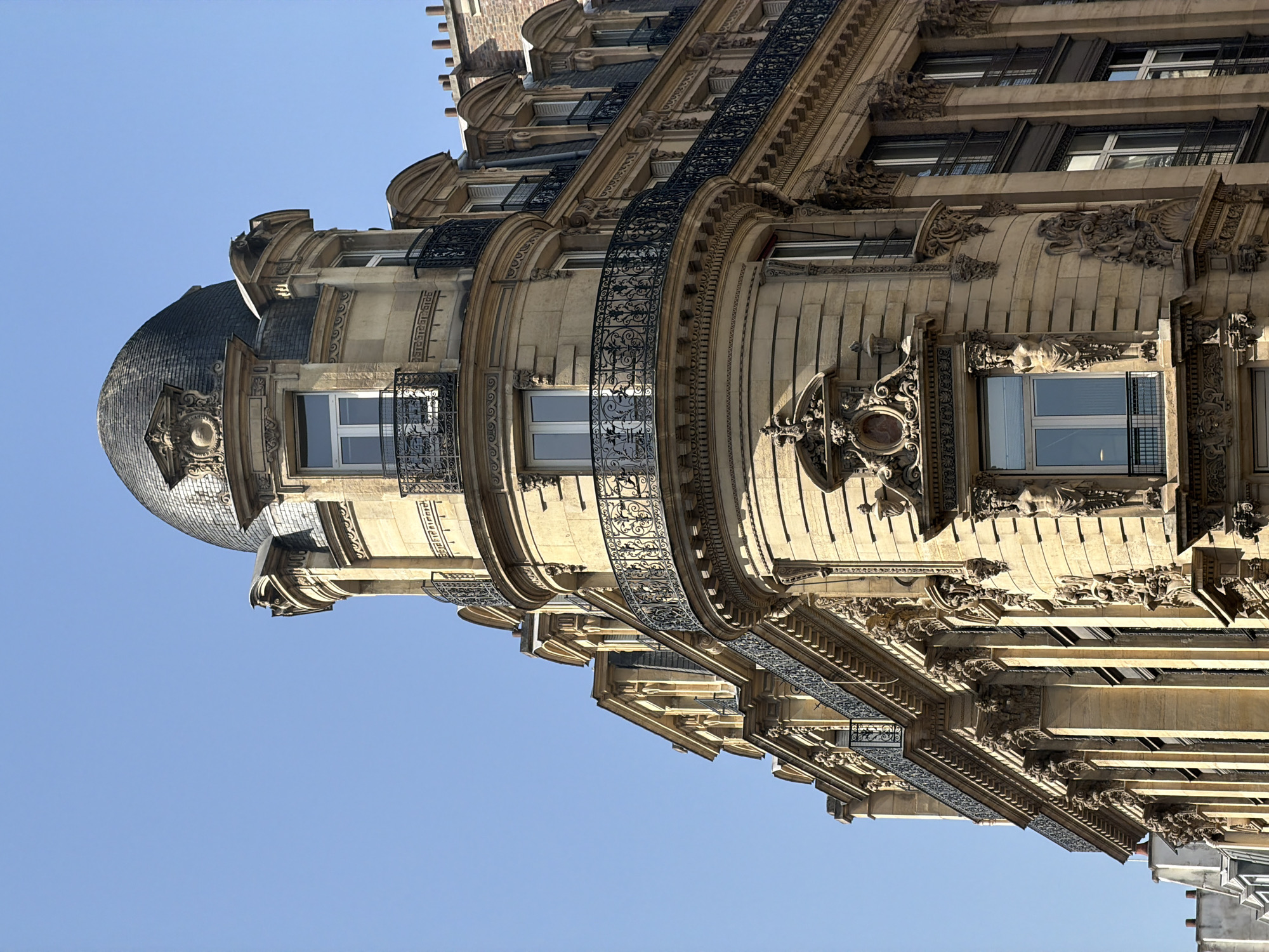 The image shows the upper portion of a Haussmannian building in Paris, France, under a clear blue sky. The building features a rounded corner with a dome roof covered in dark gray or black tiles. The facade is decorated with ornate carvings, balconies with black wrought-iron railings, and windows with white frames. The stone appears to be beige or light brown. There are no people visible in the scene.