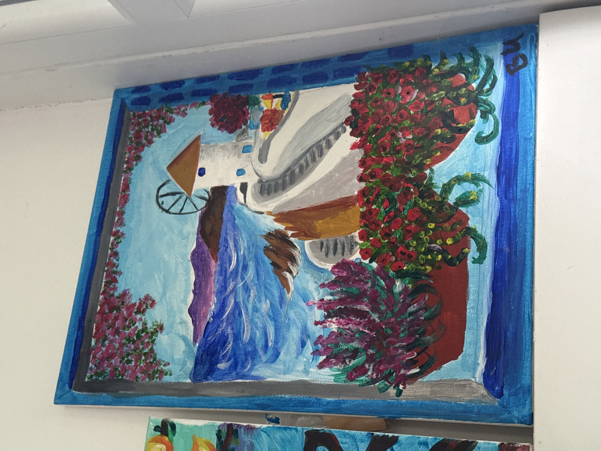 A horizontally rectangular painting is hanging on a white wall. The painting is framed in a blue border with a darker blue accent pattern. The painting depicts a stylized, rotated view of what appears to be the city of Plovdiv, Bulgaria, seen through a window or balcony. On the right, two potted plants with red and purple flowers sit, filling the majority of the space. Through the opening, a blue background suggests sky. The painting also features abstract representations of a building, a wheel and a waterfall-like flow with a small red flower detailing along a blue outline. The overall style of the painting is amateur or child-like, using flat colors and simple shapes. Another painting can be seen partially beneath this one.