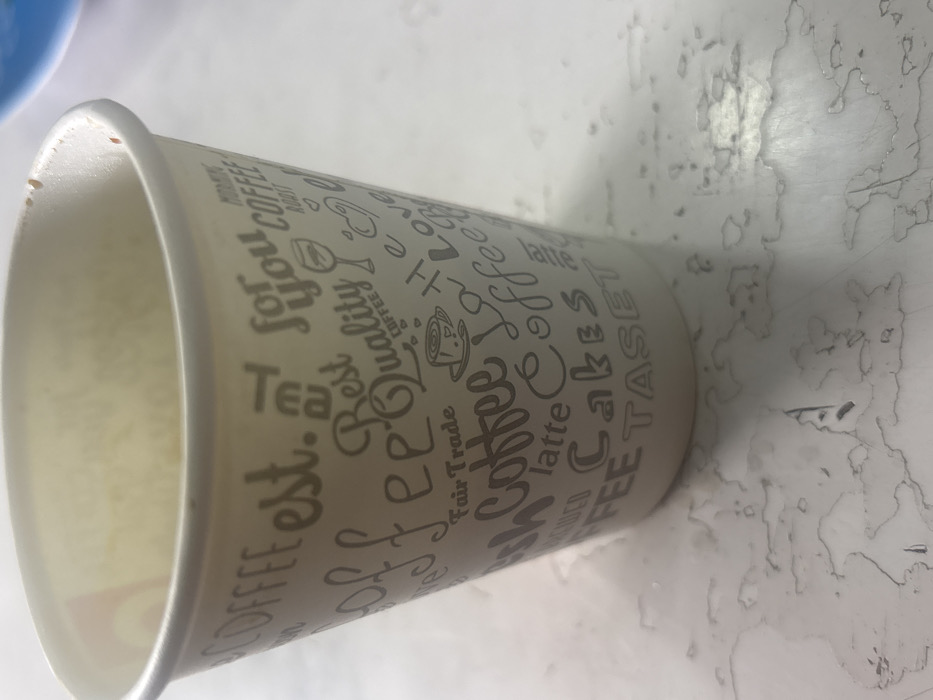 A light-toned, disposable paper cup is positioned upright on a flat, light-colored surface in the city of Lehvaz, Armenia. The cup's exterior is adorned with grey text, including words such as "COFFEE," "est.," "TEA," "for you," "Best Quality," "Fair Trade," "Latte," "Cakes," "TASET," "ROAST," and "Love," alongside small illustrations of a coffee cup, a wine glass, and a heart. Minimal brown residue is visible along the interior rim of the cup. The surrounding surface is marked by numerous irregular, dried liquid stains, appearing light brown to greyish in color, extending outwards from the cup's base. A blurred blue object is partially discernible in the upper right portion of the background. No individuals are present within the frame.