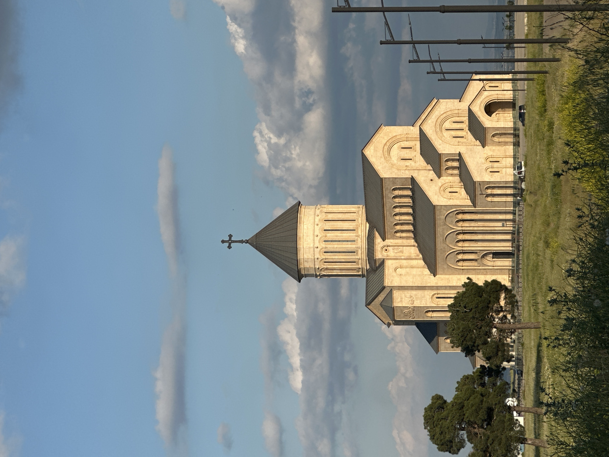 The Holy Trinity Cathedral of Tbilisi, also known as Sameba Cathedral, stands prominently against a backdrop of a partially cloudy blue sky. The massive stone structure, bathed in warm light, features multiple domes and intricate architectural details. A cross crowns the central, largest dome. In the foreground, a grassy field with wildflowers and scattered trees leads to the cathedral. Several modern streetlights line the right side of the image. Two vehicles are visible near the cathedral entrance, one white and one dark. The scene appears to be an exterior shot of a religious landmark in Tbilisi, Georgia.