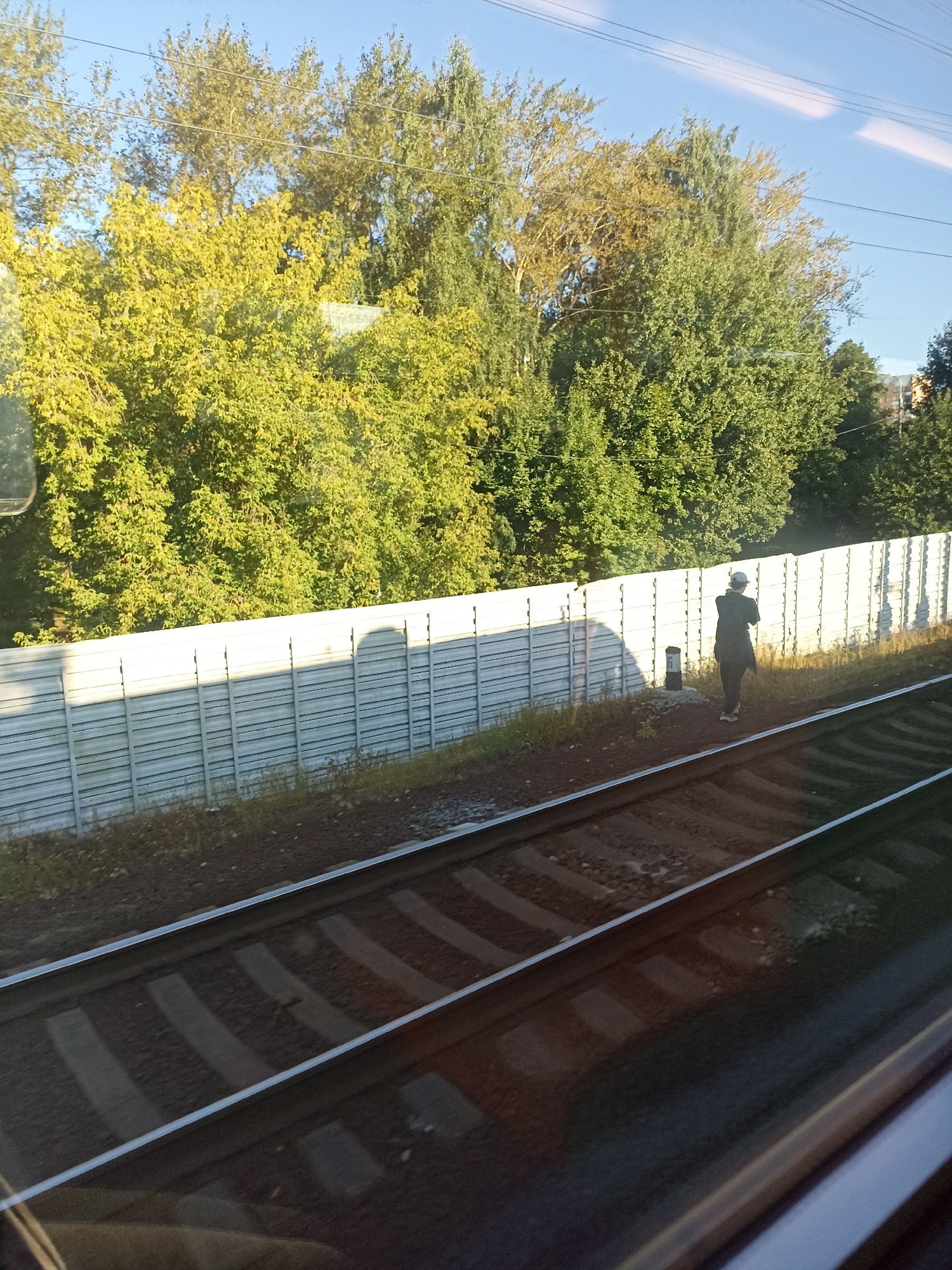 The image depicts a person standing on a dirt path beside train tracks, facing away from the camera. The person is wearing dark clothing and a white cap. A white noise barrier runs parallel to the tracks, with trees and foliage visible behind it. A small black pole with a white number "6" is situated near the person. The scene is illuminated by sunlight, casting shadows. The environment suggests an outdoor setting, likely in a suburban or industrial area near a railway line. The presence of train tracks and the noise barrier indicate a transportation corridor. The trees suggest a temperate climate.