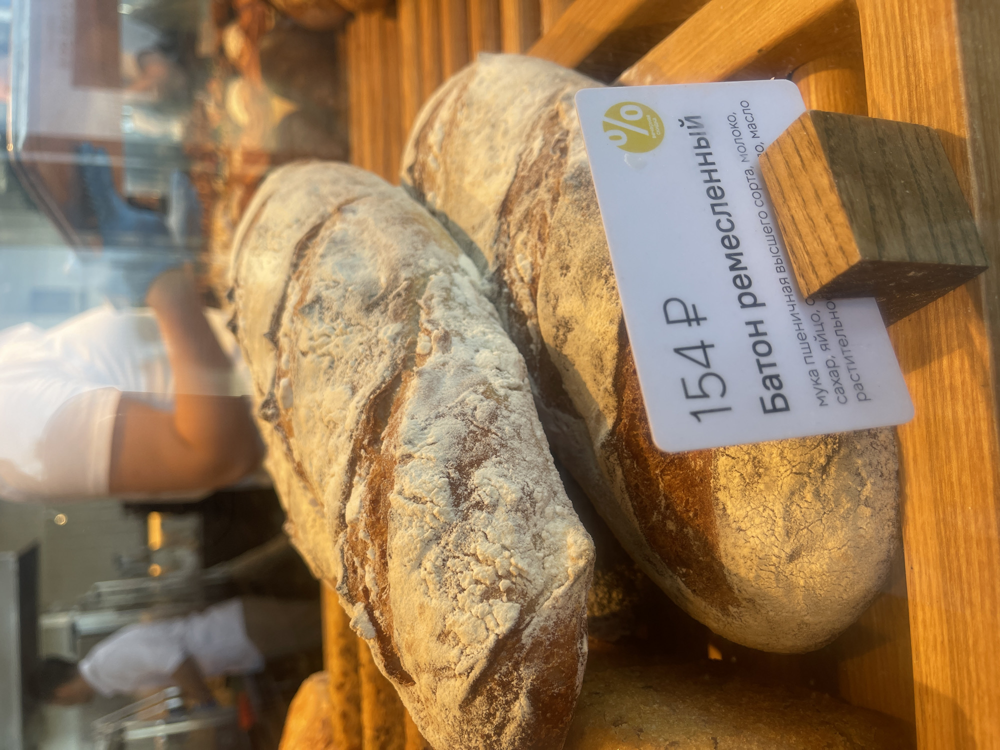 The image shows two loaves of artisan bread on display in a bakery. The bread is dusted with flour and has a rustic, golden-brown crust. A price tag is visible, indicating "154 ₽" and "Батон ремесленный" (Artisan Baton). Below this, the ingredients are listed: "мука пшеничная высшего сорта, молоко, сахар, яйцо, сливочное масло, растительно" (high-grade wheat flour, milk, sugar, egg, butter, vegetable oil). The bakery appears to be located in Zelenogradsk, Russia, based on the visible Cyrillic text and the general context of a bakery display. In the blurred background, a person wearing a white shirt is partially visible, suggesting a retail environment. The lighting is warm, highlighting the texture of the bread.