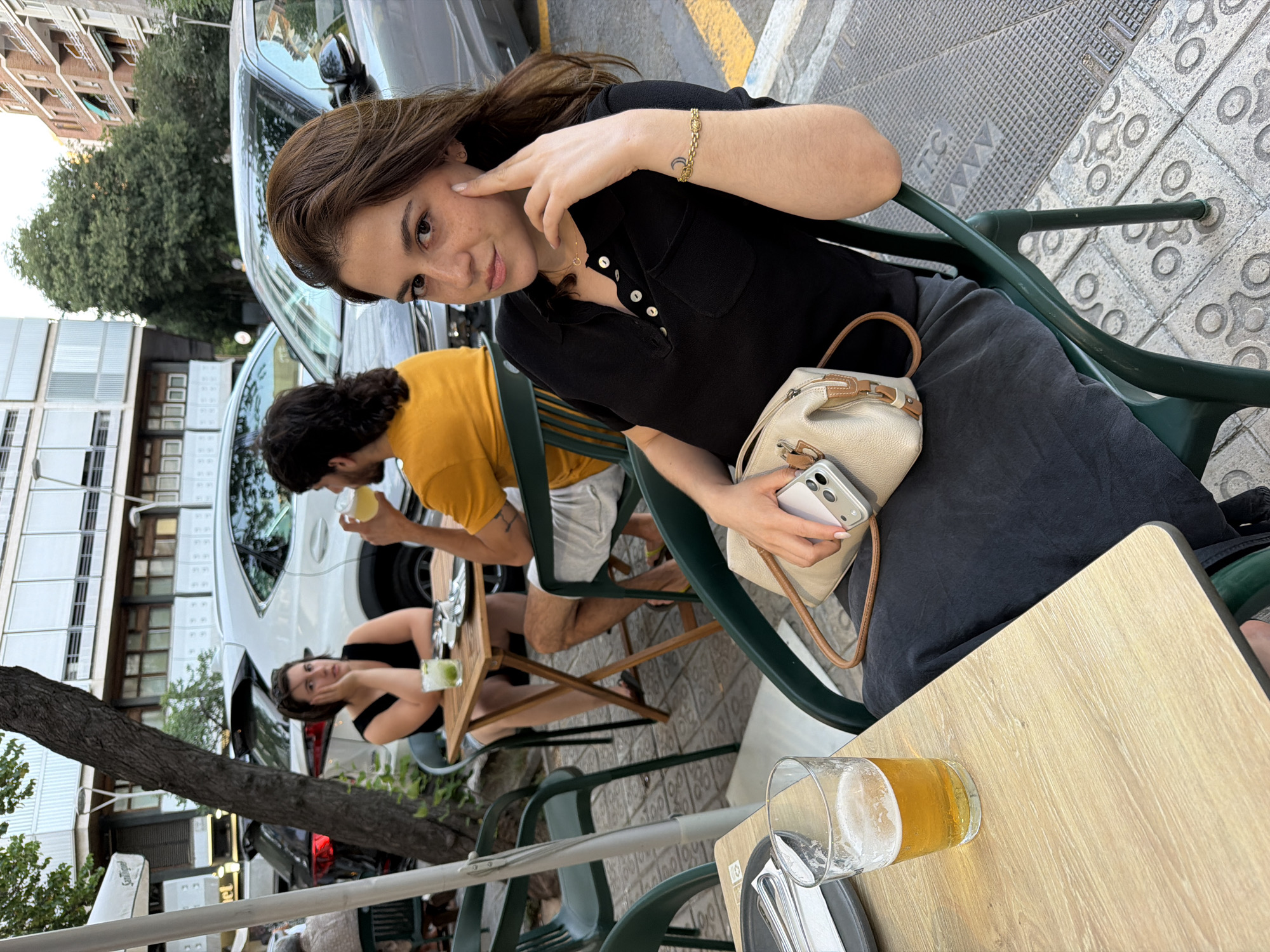 A young woman with brown hair and freckles, wearing a black polo shirt and a dark skirt, sits at an outdoor cafe table. She rests her left hand on her cheek and her right hand holds a smartphone. Next to her, a glass of beer sits on the light wood table. In the background, a man in a yellow shirt and a woman in a black dress are also seated at a table. Cars are parked on the street behind them. The scene appears to be on a sidewalk in a city, possibly Barcelona, Spain, given the architectural style of the buildings and the patterned pavement.