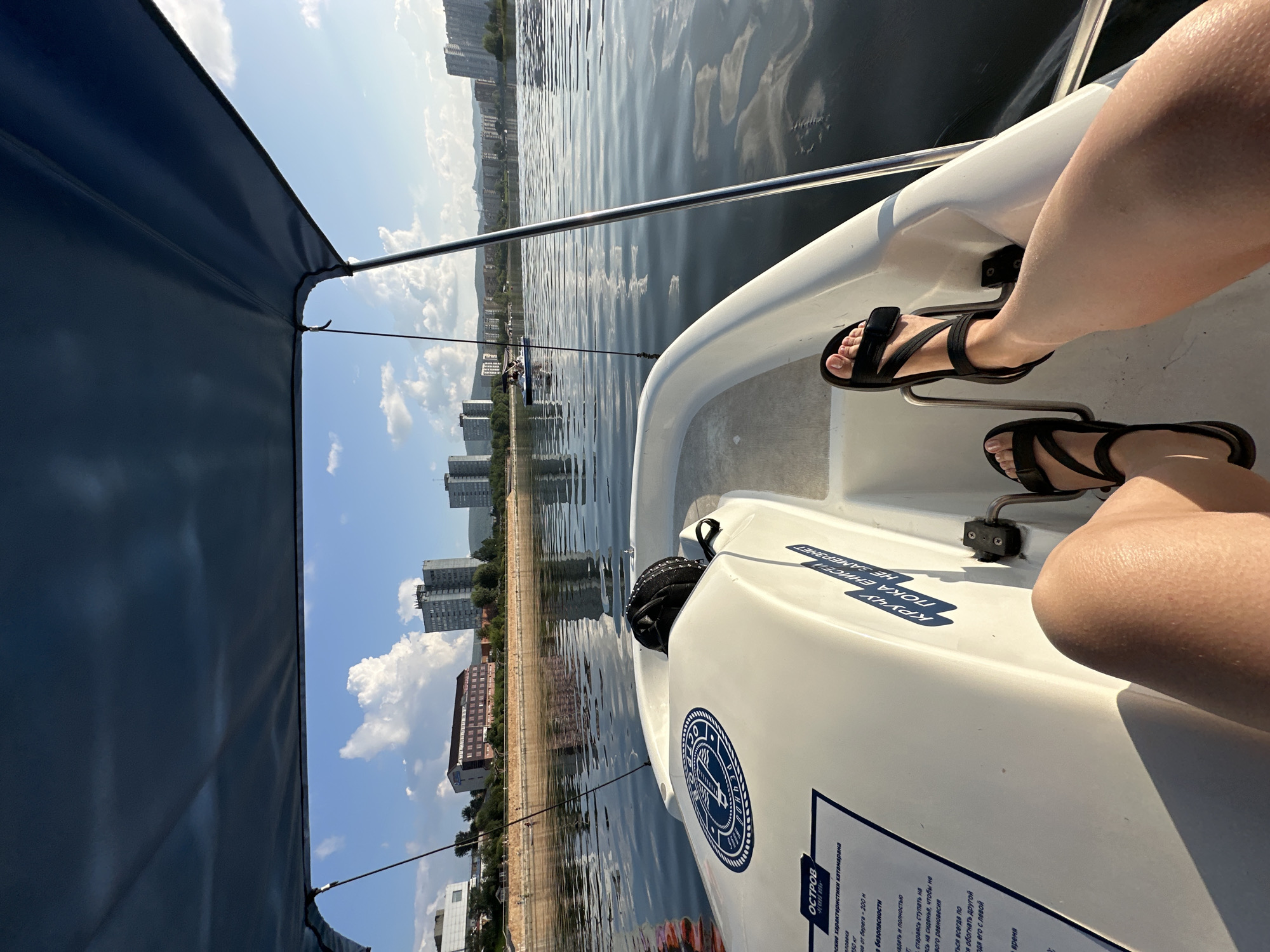 In the foreground of the image, a person's legs and feet are visible from the knees down, wearing black sandals. The person is seated in a white pedal boat on a body of water. The boat has a dark blue canopy overhead. In the background, a city skyline with several tall buildings is visible across the water. Trees and a waterfront promenade are also present. The sky is blue with scattered white clouds, and the water reflects the sky and buildings. The overall scene suggests a recreational activity on a lake or river within an urban area.