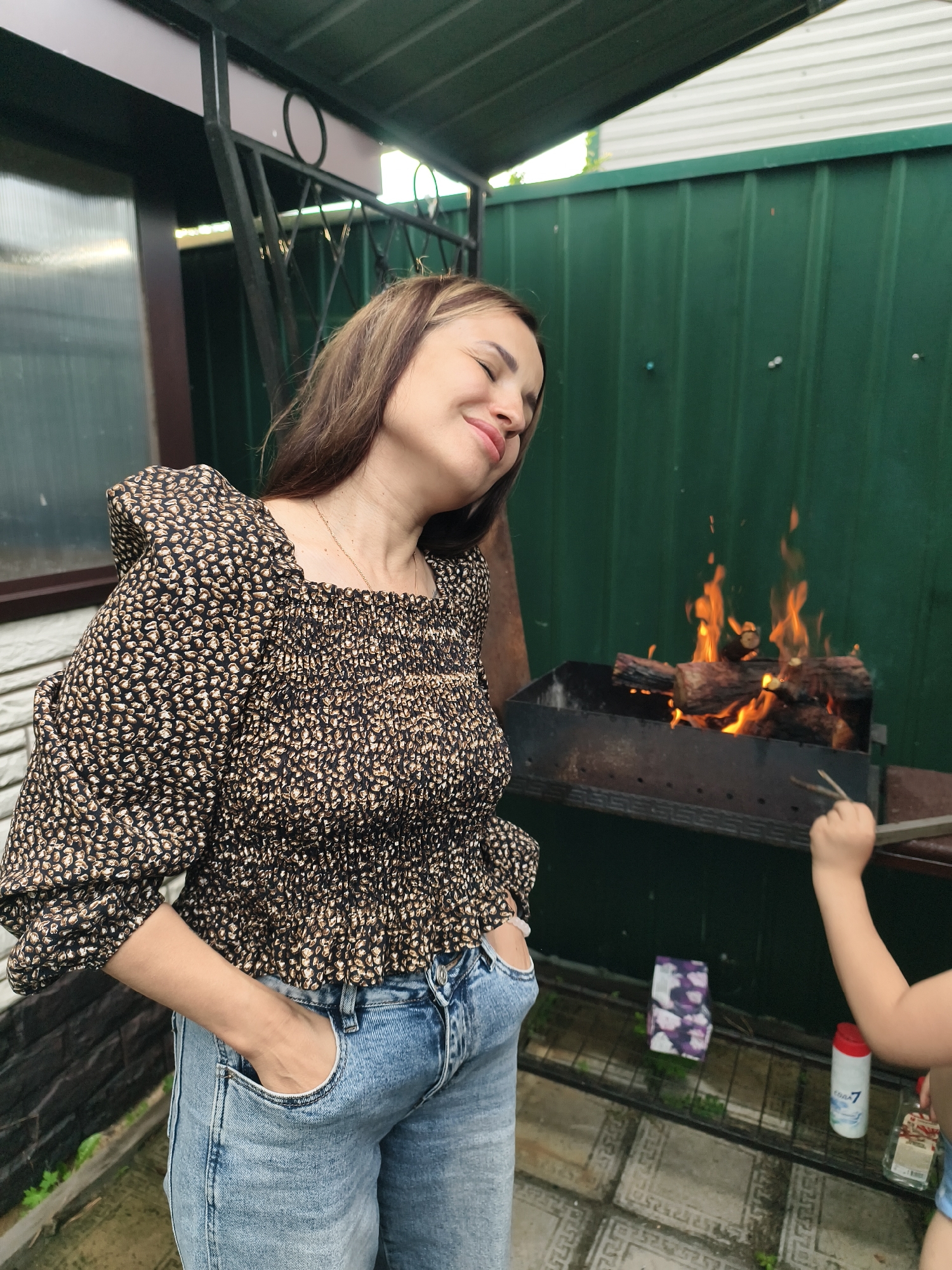 The image shows a woman standing in front of a barbecue grill with a fire burning. She is wearing a patterned top and blue jeans and has her hands in her pockets. The woman has her eyes closed and a relaxed expression on her face. To the right of the grill, a child's hand is visible, holding a barbecue utensil. The background consists of a green fence and a covered patio area. The overall scene suggests a casual outdoor gathering. The time of day appears to be daytime, possibly late afternoon or early evening, given the lighting.