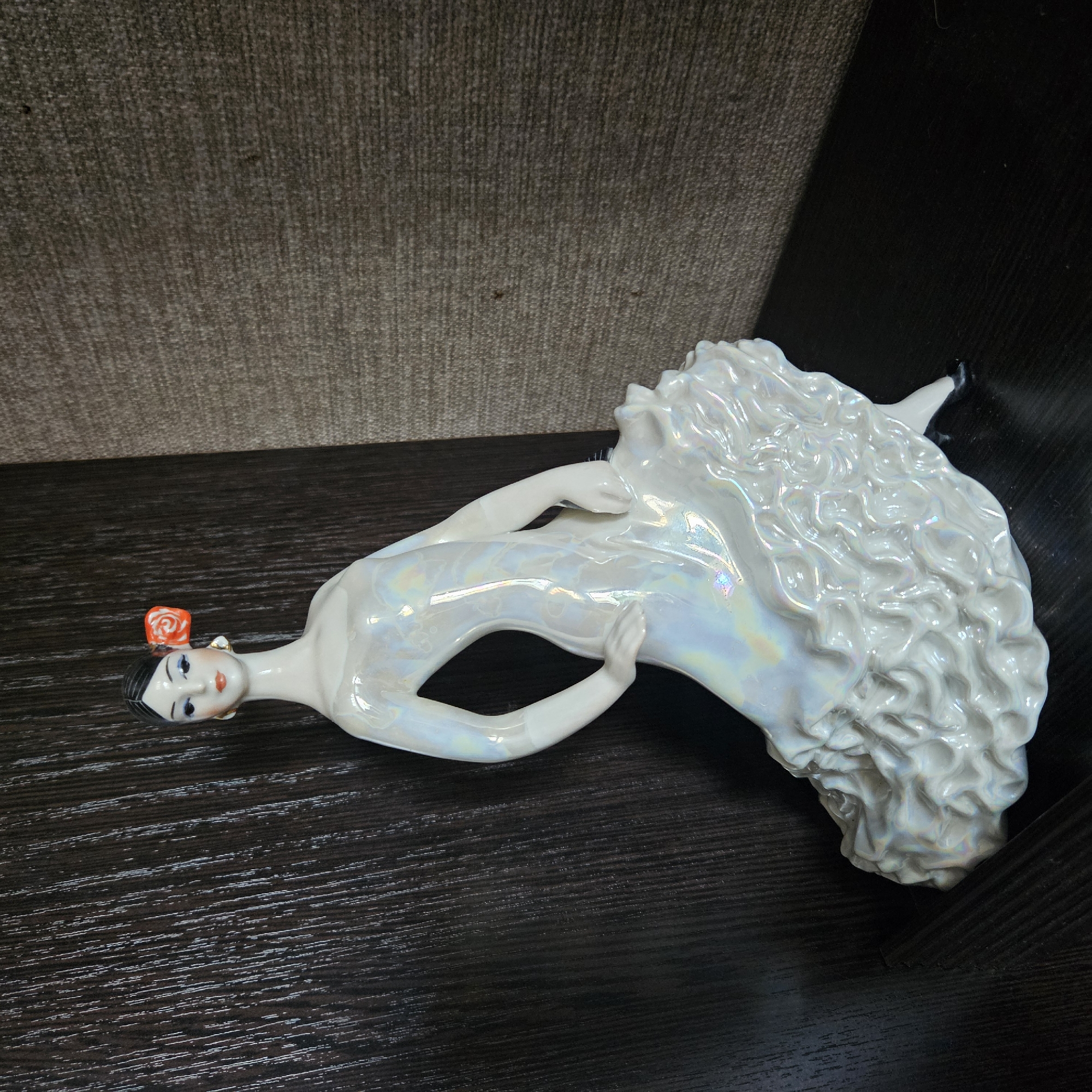 A porcelain figurine of a flamenco dancer stands on a dark, polished surface. The dancer is depicted in a dynamic pose, with her body tilted and her skirt fanned out. She wears a shimmering white dress with iridescent highlights, and her hair is styled up with an orange rose tucked behind her ear. The background consists of a dark, wood-grained surface on the left and a textured, gray material on the right, suggesting an indoor shelf or display area. No specific location in Chelyabinsk, Russia, can be identified from this image.