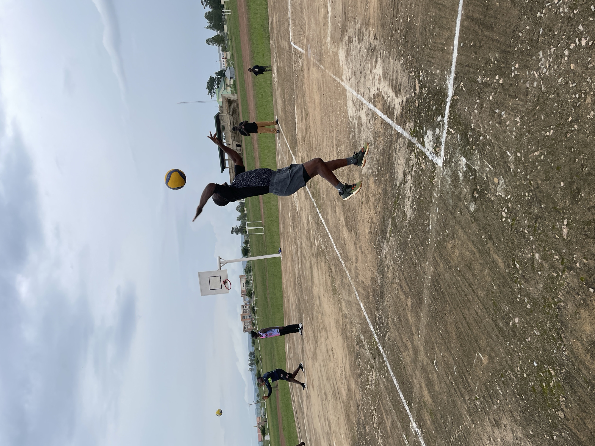 In Durumi, Nigeria, a group of young men are playing volleyball on an outdoor court. The court is made of concrete with white lines marking boundaries. In the foreground, one player is in mid-air, about to serve the ball, which is yellow and blue. He is wearing a black t-shirt with a patterned design and grey shorts. Other players are visible in the background, one near a basketball hoop, and others on the court. The sky is overcast with grey clouds, and the surrounding area appears to be a sports field with some buildings and trees in the distance.