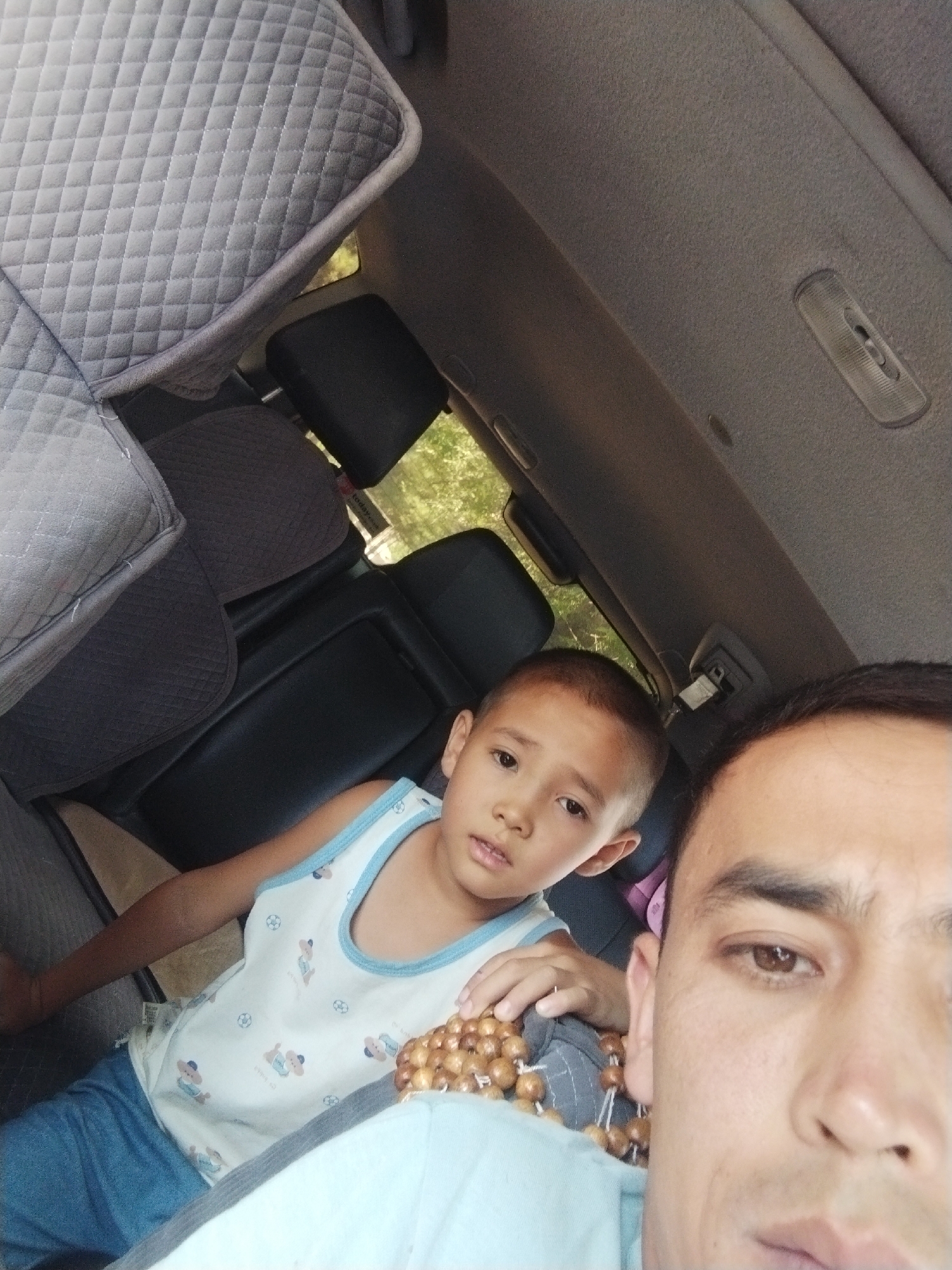 This is a close-up selfie taken from inside a car, showing two individuals: a child and an adult. The child, positioned in the mid-ground, is looking towards the camera with a slightly furrowed brow. They are wearing a white tank top with a light blue trim, adorned with a pattern of cartoon bears and soccer balls. The child's legs are covered by blue pants. A string of brown beads rests on the child's chest. In the foreground, the right side of the adult's face is visible, with their right eye looking towards the camera. The adult has dark hair and brown eyes. The background of the image reveals the interior of the car, including quilted seat covers and headrests. Through the car windows, greenery and sunlight suggest an outdoor setting, possibly during the day. The specific location in Mailuu-Suu City, Kyrgyzstan, cannot be determined from the visual information provided in the image.