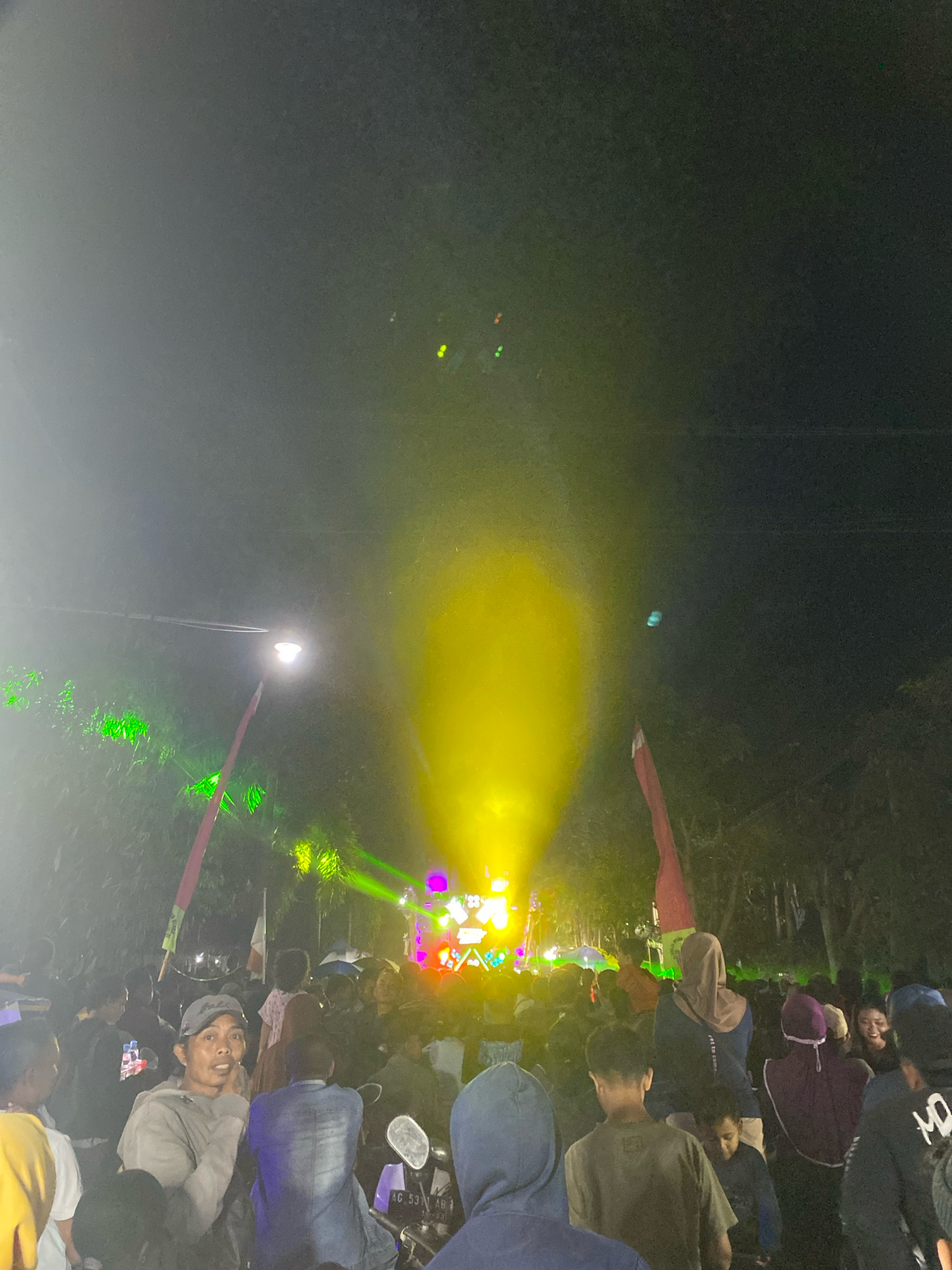 The image depicts a nighttime outdoor event in Branggahan, Indonesia, characterized by vibrant stage lighting and a large gathering of people. A prominent yellow light beam emanates from the background, illuminating the smoky atmosphere and creating a dramatic visual effect. Surrounding this central illumination are various colored lights, including green and pink, contributing to a festive ambiance.
In the foreground, a crowd of individuals are visible, many of whom are looking towards the source of the lights, suggesting a performance or event is taking place. One man in the mid-left of the frame, wearing a gray cap and sweatshirt, is looking directly at the camera with his hand resting on his chin, appearing to be a spectator. Other individuals are seen from behind, engaged with the event.
The environment suggests a public space, possibly a park or an open field, with trees and foliage visible in the background, partially obscured by the lighting and the crowd. A streetlamp on a pole is also present, adding to the overall illumination of the scene. The presence of a motorcycle in the lower right corner, with visible license plate "AG 5371 AB", indicates a mode of transportation common in the area and grounds the scene geographically within Indonesia. The overall impression is one of a lively community gathering, possibly a concert or festival, taking place after dark.