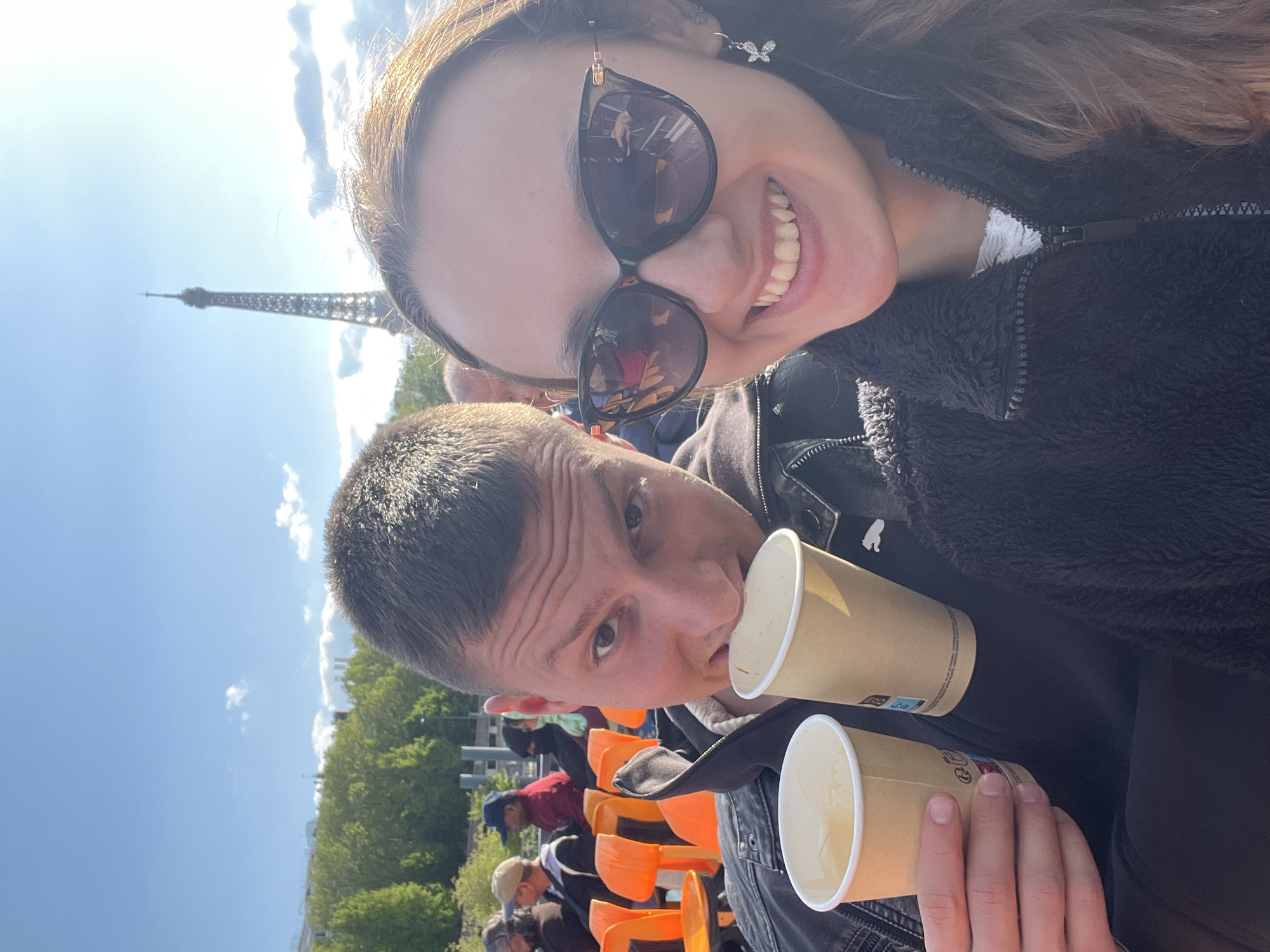 This media file is a bright, sunlit selfie featuring a young woman and a young man, taken outdoors in Paris, France, with the Eiffel Tower clearly visible in the background.
The young woman, positioned in the upper right, is smiling broadly at the camera. She wears large, dark cat-eye sunglasses that reflect a blurry image of the surroundings. Her long, dark hair cascades around her face, and she has a distinctive butterfly-shaped earring on her right ear. She is dressed in a dark, textured, zippered jacket.
The young man is in the lower half of the frame, looking towards the camera with a neutral, slightly pensive expression. He has short, dark hair and wears a dark jacket. He holds two light brown paper cups; one is raised close to his mouth, and the other is held lower.
In the background, the iconic Eiffel Tower stands tall against a clear, vibrant blue sky, dotted with a few scattered white clouds. This prominent landmark firmly establishes the location as Paris. Below and to the left of the man, several other people can be seen seated in rows of bright orange chairs, suggesting an outdoor public space or viewing area, possibly a cafe or observation deck. Green trees are also visible in the distance. The bright natural light indicates it is daytime. There is no clearly legible text on the paper cups.