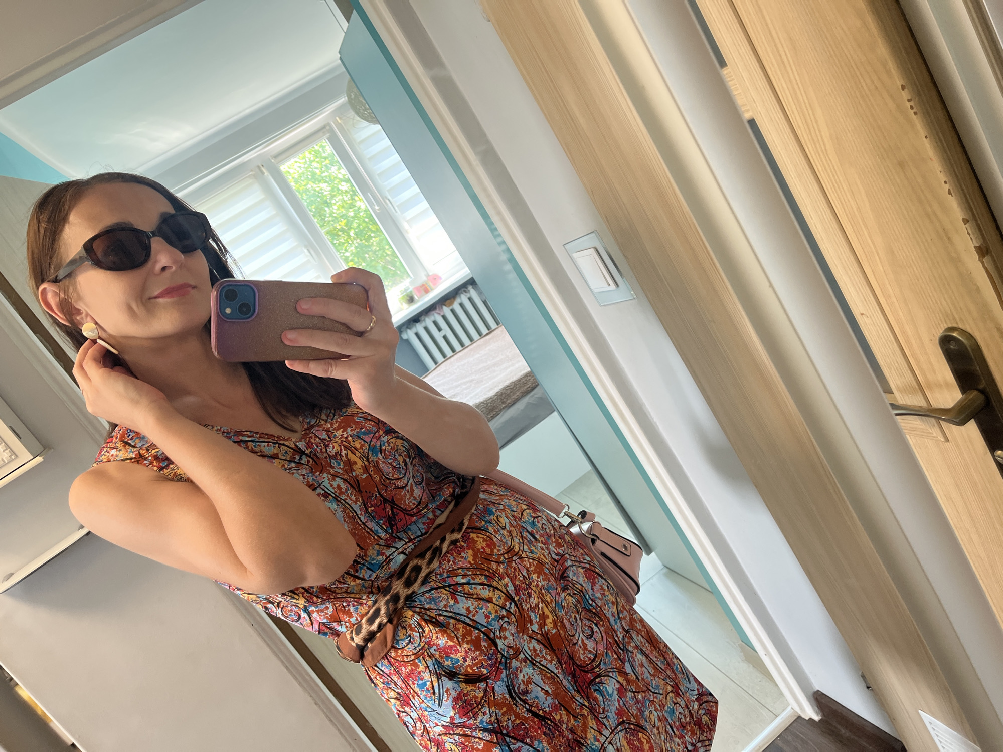 In this mirror selfie, a woman with brown hair and sunglasses is taking a picture of herself. She is wearing a colorful, abstract print dress with a leopard print belt. She has a pink purse slung over her shoulder. The background shows a room with a window that lets in natural light, suggesting it is daytime. The room appears to be a bedroom or dressing area, with a radiator visible beneath the window. The woman is touching her ear, possibly adjusting an earring. She is smiling slightly, conveying a confident and pleasant mood. The overall impression is of a casual, everyday moment captured in a domestic setting.