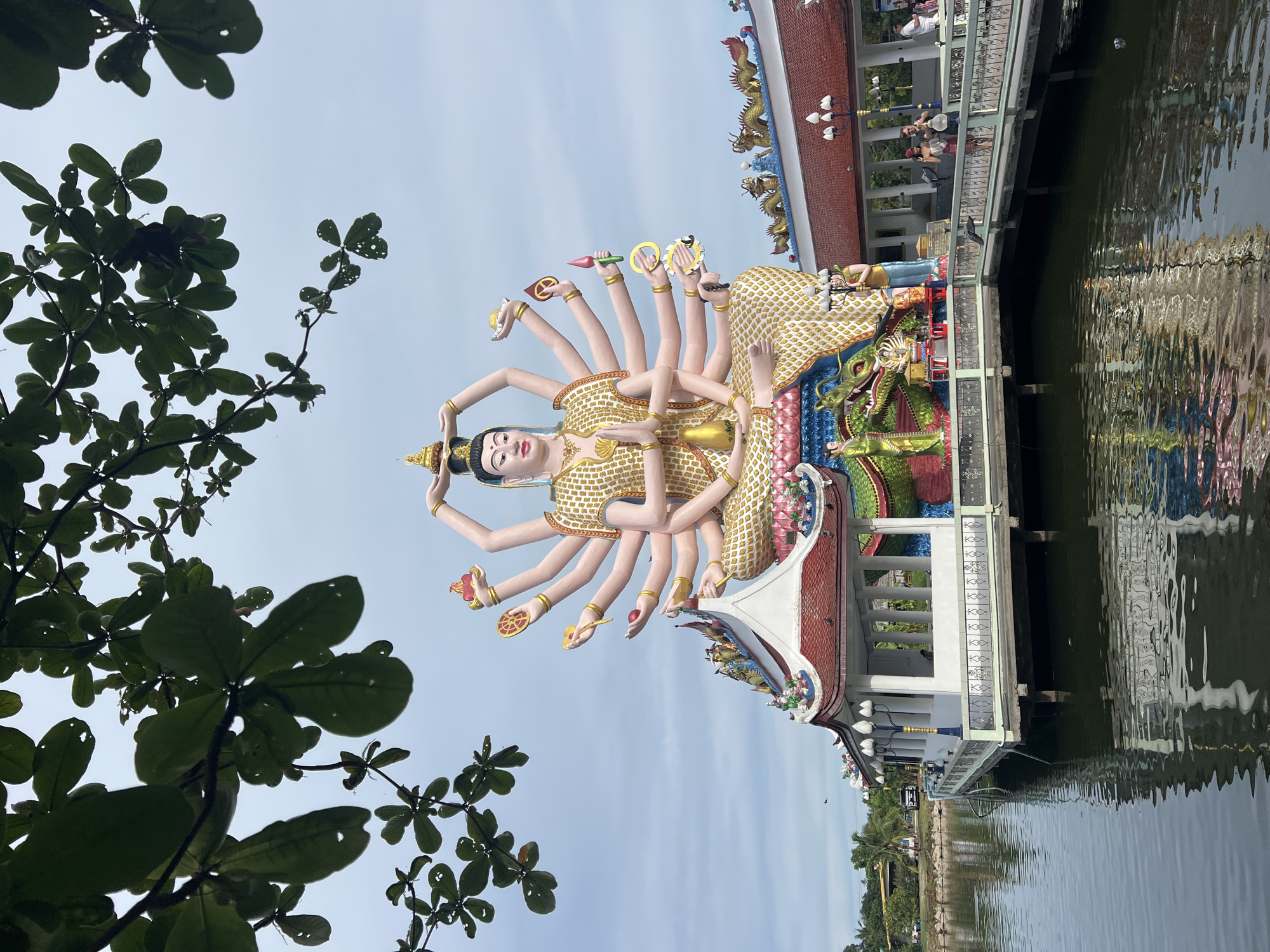 In Baan Plai Laem, Thailand, a multi-armed statue of a deity sits majestically on a lotus pedestal, its serene face gazing forward. The statue is adorned in a patterned golden garment, with each hand holding a symbolic object, such as a wheel, a jewel, or a lotus flower. The deity is positioned in front of an elaborate temple complex, featuring multi-tiered roofs with intricate dragon and floral motifs. A walkway with ornate railings extends over a tranquil body of water, where reflections of the temple and statue are visible. Several people are present, some walking on the walkway and others observing the surroundings. Lush green trees with large leaves frame the upper portion of the image, contrasting with the vibrant colors of the temple architecture. The sky is overcast with diffused light.