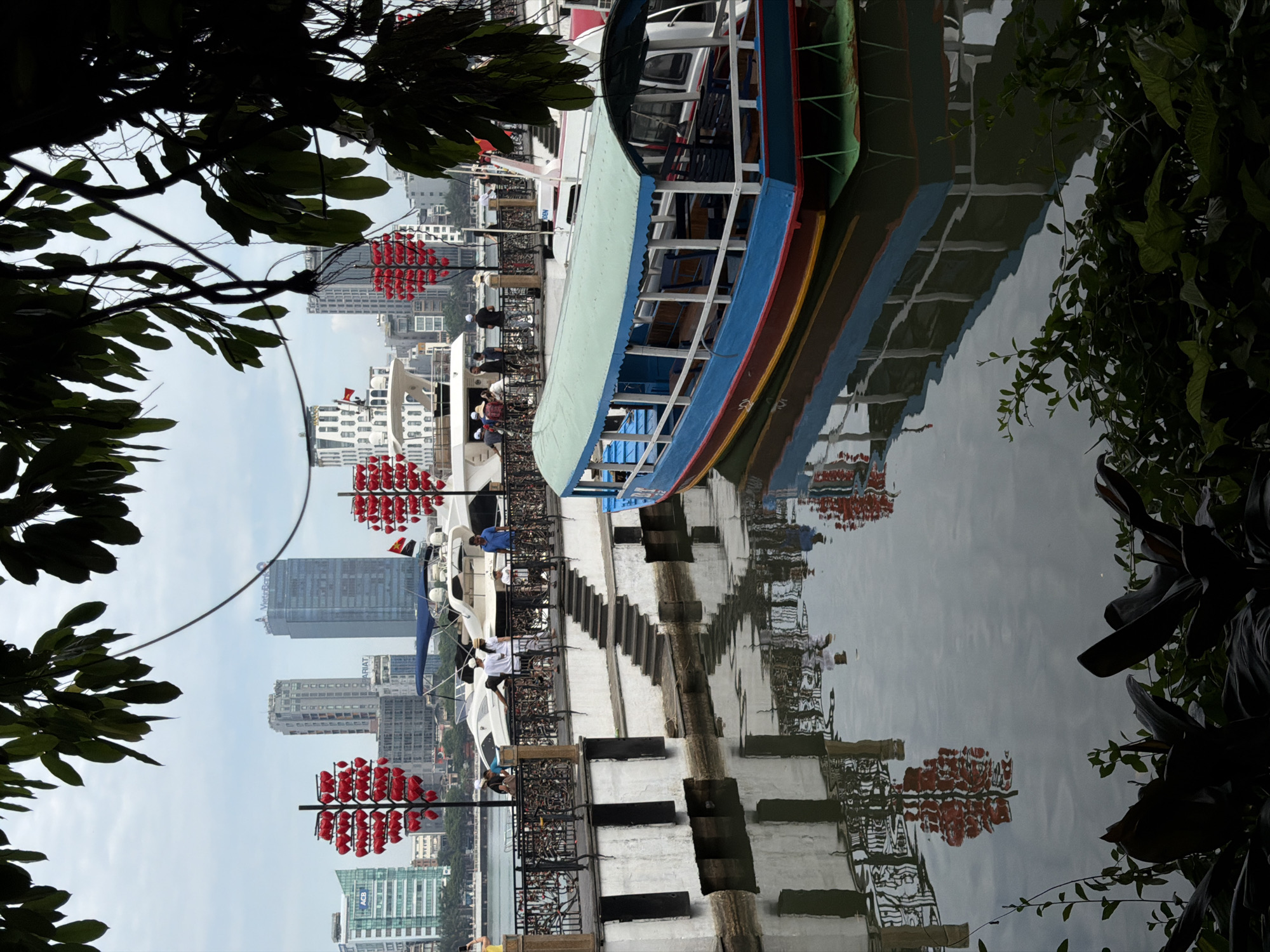 The image depicts a waterfront scene in the city of Đà Nẵng, Vietnam, likely along the Han River. In the foreground, lush green foliage partially obscures the view. The middle ground features a walkway with decorative red heart-shaped lanterns hanging from poles. Several people are visible on the walkway, some standing and others appearing to walk. Bicycles are also present, secured to railings. A boat with a blue and red hull and a light green canopy is docked in the water to the right, its reflection visible on the calm surface. In the background, several modern high-rise buildings line the city skyline, including structures with the "ACB" and "Vinhomes" logos. A large white yacht is also visible docked near the walkway. The sky is overcast, suggesting a cloudy day. The overall impression is of a vibrant urban setting with a blend of natural elements and man-made structures.