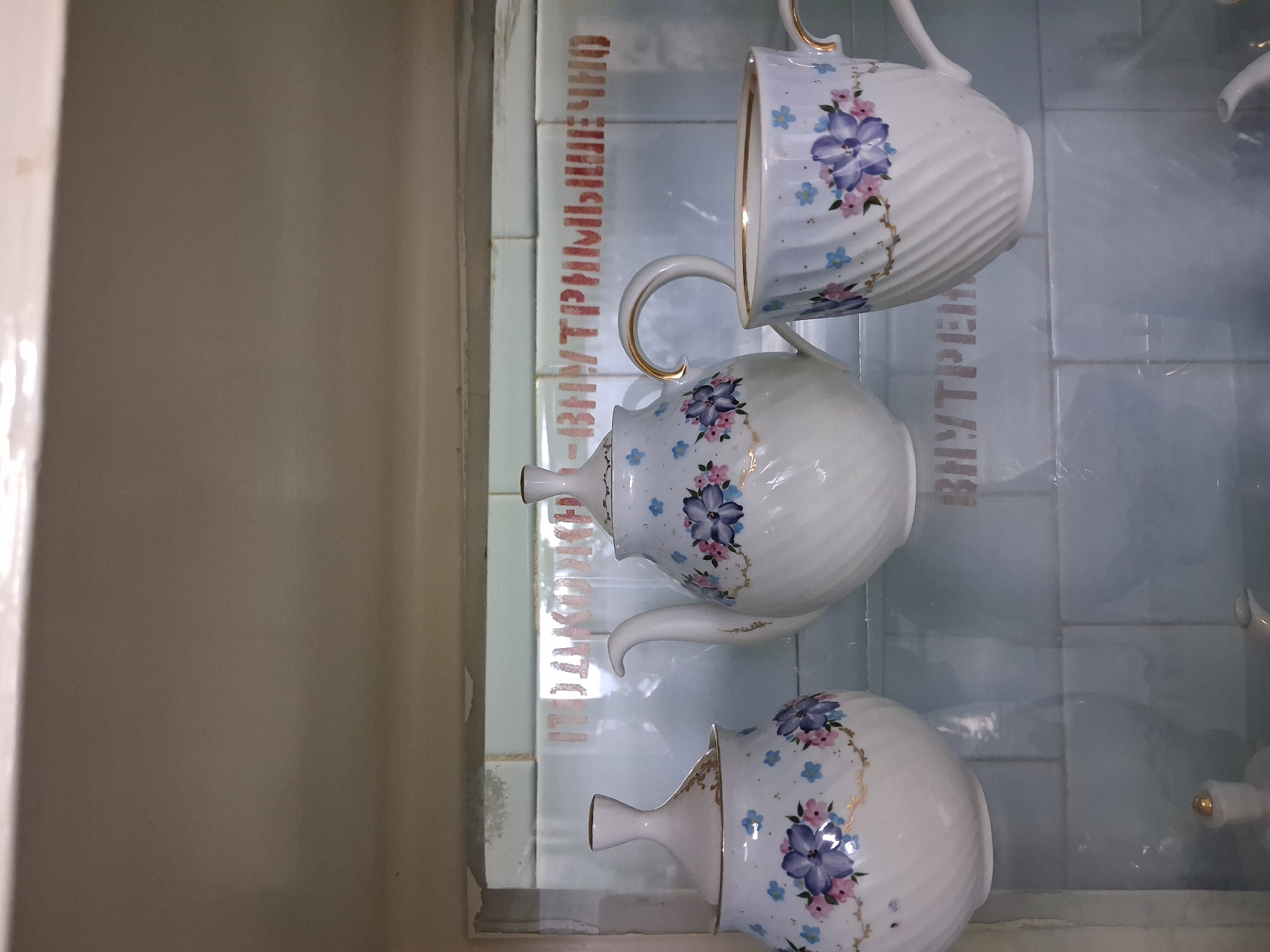 The image depicts a white porcelain tea set with blue and pink floral patterns displayed inside a cabinet. The tea set consists of a teapot, a sugar bowl with a lid, and a teacup. The cabinet appears to be part of a kitchen or dining area, with light-colored tiled walls in the background. Red Cyrillic text, partially obscured, is visible on the tiled wall behind the tea set. This scene is likely located in a private residence in Sysert, Russia, given the Cyrillic text.