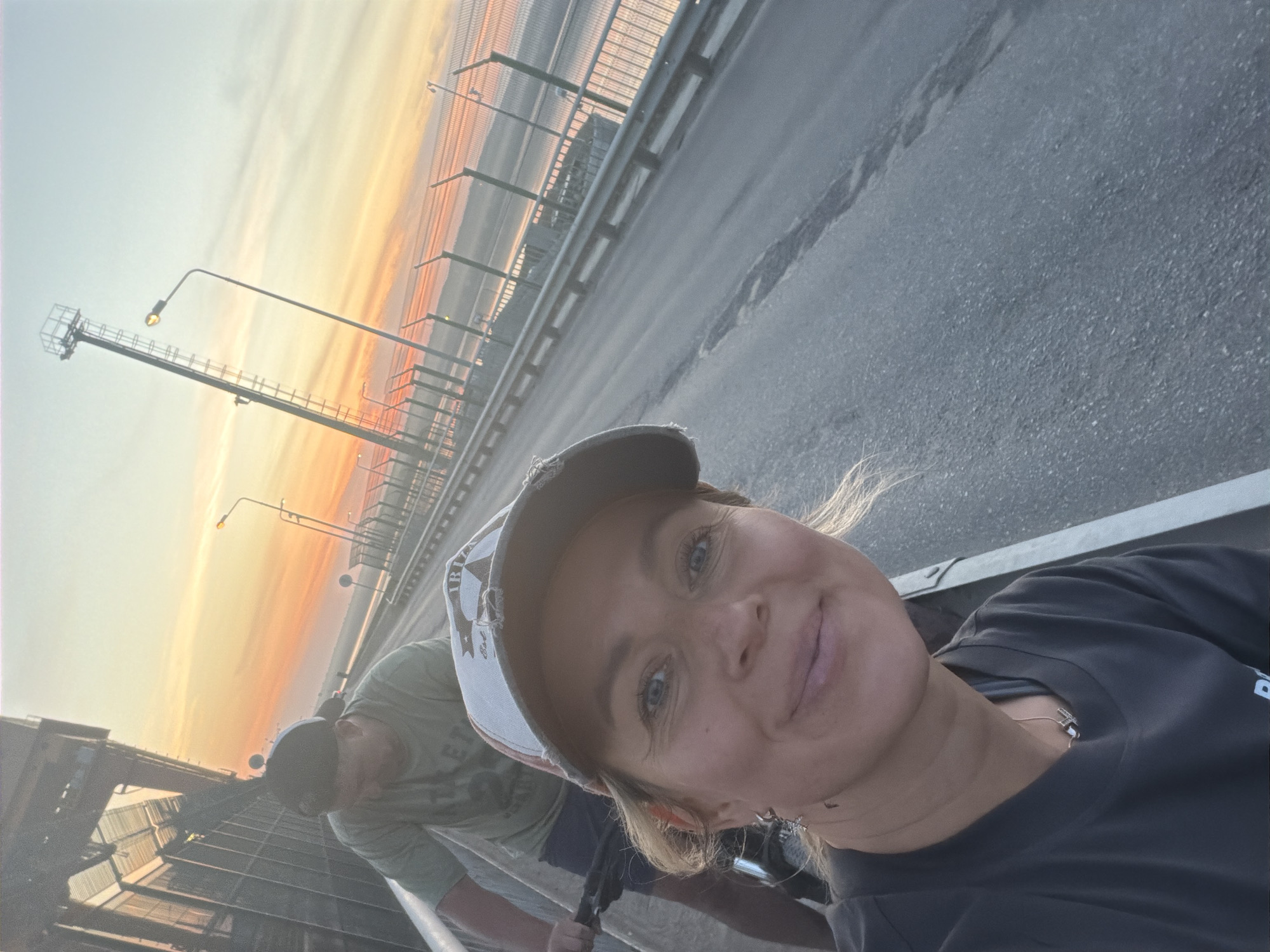 A selfie-style photograph shows a woman wearing a dark t-shirt and a baseball cap, with a person in the background wearing a green t-shirt and baseball cap. The background appears to be a bridge at sunset or sunrise, with the sky displaying orange and yellow hues. The bridge has railings and light poles visible along the side. The location is identified as being in Chaikovsky, Russia, though specific details confirming this are not visible in the image alone.
