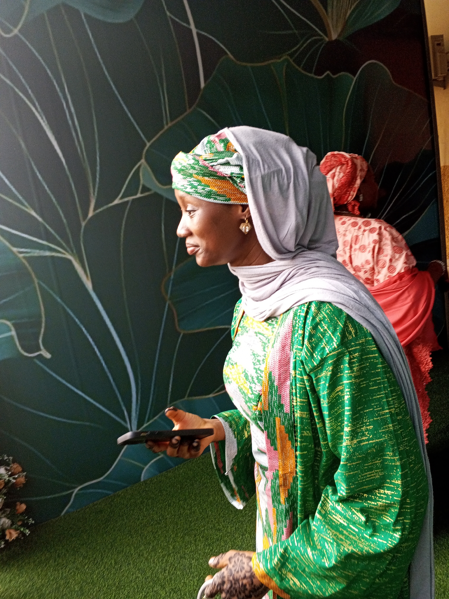 In Abuja, Nigeria, a woman dressed in a green and gold patterned kaftan and a grey hijab stands on artificial turf. She is wearing a gold heart-shaped earring. In her right hand, she holds a black smartphone. Her left hand, adorned with intricate henna designs, is visible at the bottom right of the frame. Behind her, another woman in a pink and white patterned outfit and a red head covering is partially visible. The background features a large mural of dark green leaves with white vein patterns. A bouquet of light-colored flowers is positioned in the lower left corner of the image. A utility box is mounted on the wall to the right of the mural.