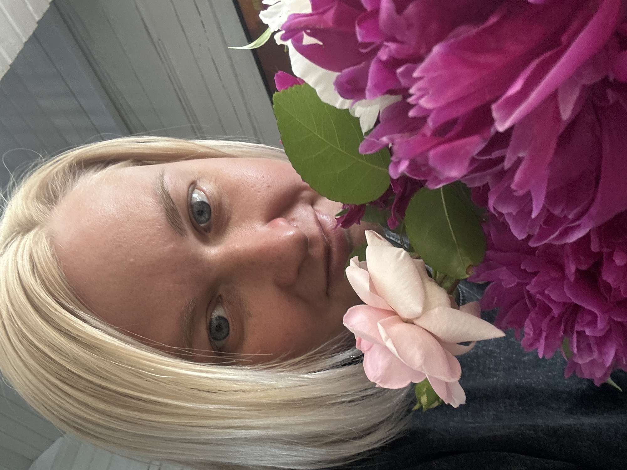 A close-up, rotated selfie shows a person with fair skin and blonde hair looking directly at the camera. Their left eye, a piercing blue, is clearly visible, as is a portion of their nose and lips. The person appears to be holding a bouquet of flowers, with large, vibrant pink peonies and a soft pink rose prominently featured in the foreground. Green leaves from the bouquet also add color to the composition. The background is blurred but suggests an outdoor setting, possibly a porch or garden, with horizontal wooden or siding elements visible. The lighting indicates it is daytime, and the overall mood is relaxed and personal. There is no visible text in the image.