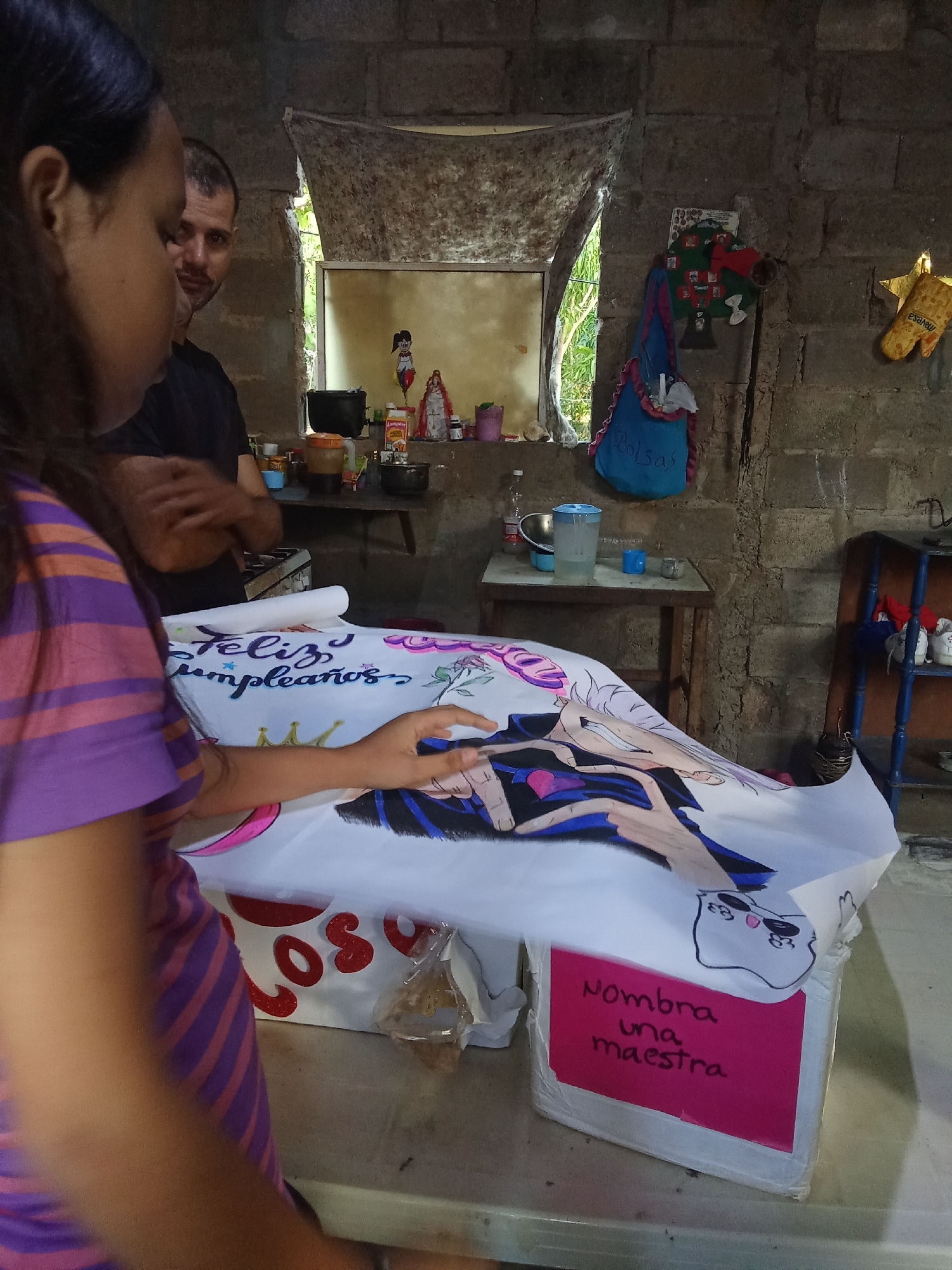 The image shows an indoor scene in what appears to be a residence in Juan Griego, Venezuela.
A woman wearing a striped purple shirt is in the foreground. She is looking down and pointing with her right hand towards a large white banner or poster that is lying on a surface, possibly a table. The banner has "Feliz Cumpleaños" (Happy Birthday) written on it and an animated drawing. A pink sign on the side has "Nombra una maestra" (Name a Teacher).
Behind the woman is a man with short dark hair and a beard. He appears to be observing the scene.
The room has exposed brick walls and a window covered with a curtain. A table is against the wall. Household items such as bottles, jars, and a water pitcher are visible on the table and in the background. A blue bag with the word "Bolsas" (bags) is hanging on the wall next to the window. A photo tree is hanging on the wall.