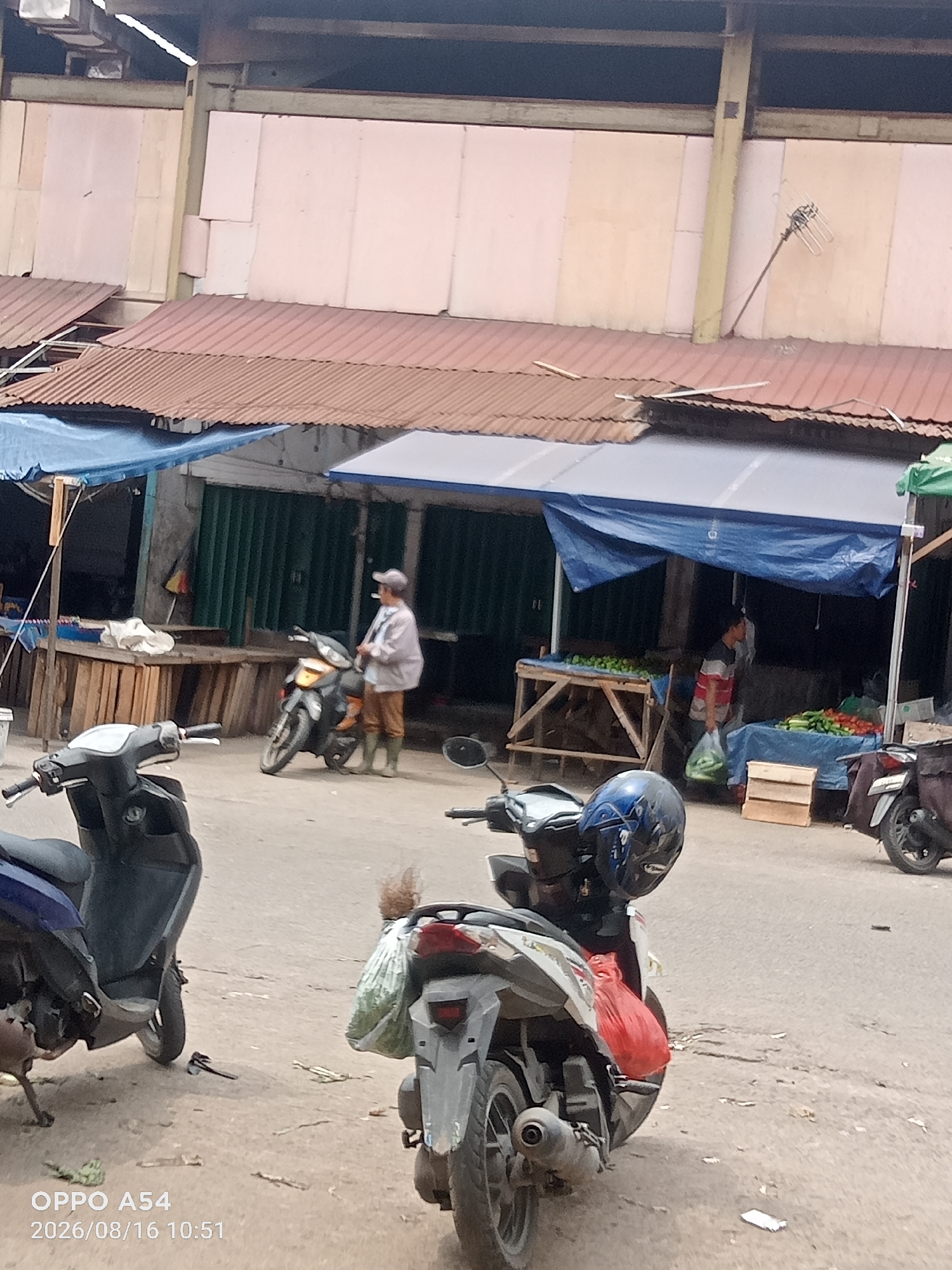 The image captures an outdoor market scene in Jambi, Indonesia, with several motorcycles parked on a paved surface. In the foreground, a dark-colored scooter is partially visible on the left. Further back, a person wearing a light-colored jacket, brown pants, and green boots stands next to a parked motorcycle, appearing to be smoking a cigarette. Behind the person, a market stall covered with a blue tarp displays green produce on wooden tables. Another stall to the right, also with a blue tarp, showcases a variety of vegetables, including cucumbers and tomatoes. In the background, market structures with corrugated metal roofs are evident, and a beige wall with some pinkish panels forms the backdrop. The overall environment suggests a bustling local market setting.