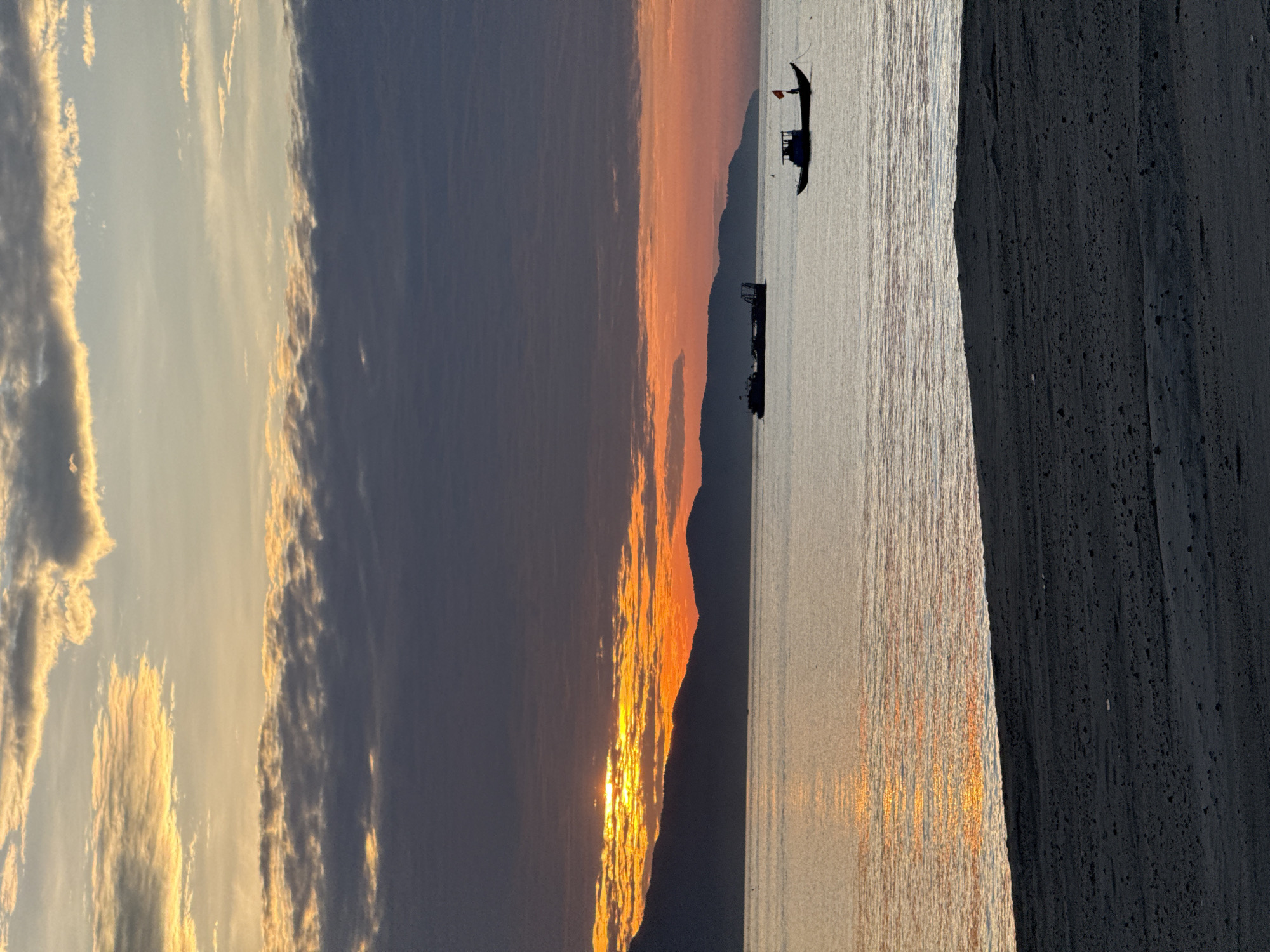 This detailed image captures a sunrise or sunset over the sea in Đà Nẵng, Vietnam. The sky dominates the upper portion, featuring a dramatic display of colors transitioning from vibrant orange and yellow near the horizon, where the sun is partially visible behind dark clouds and mountains, to lighter blues and grays higher up, with some clouds illuminated golden. A range of dark, silhouetted mountains forms the distant coastline, spanning across the middle of the frame. The calm sea reflects the sky's warm hues, exhibiting a golden-orange shimmer, particularly on the left side, with subtle ripples on the surface. Two boats are visible: a larger, darker vessel, possibly a barge or dredge, is silhouetted in the middle-ground to the left, and a smaller, traditional fishing boat is positioned to the right, also in silhouette, identifiable by its distinct red flag (likely the flag of Vietnam) raised on a pole at its stern. The foreground consists of a dark sandy beach, occupying the bottom quarter of the image, showing fine granular texture and slight undulations. No human figures are present.