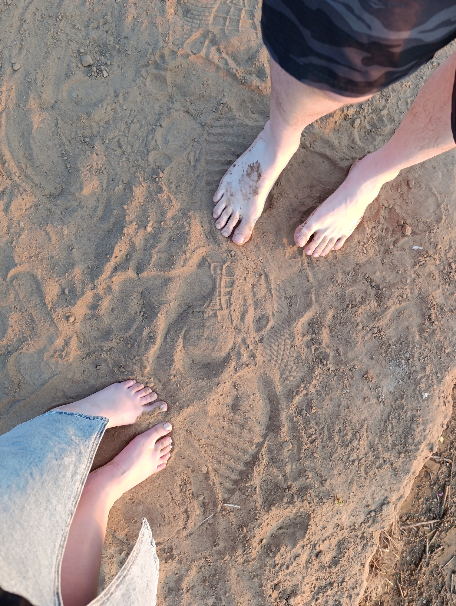 An overhead, eye-level shot shows two pairs of bare feet on sandy ground. One pair of feet, belonging to a person wearing camouflage shorts, is in the upper right quadrant. The feet are pale and appear to have some dirt or sand on them. The other pair of feet is in the lower left quadrant, belonging to a person wearing light-wash denim pants. These feet are also pale with slightly lighter toenails. There are footprints visible in the sand, suggesting people have walked there. The ground is textured with small pebbles and dry organic debris. The lighting suggests it is either early morning or late afternoon, with warm light casting shadows.