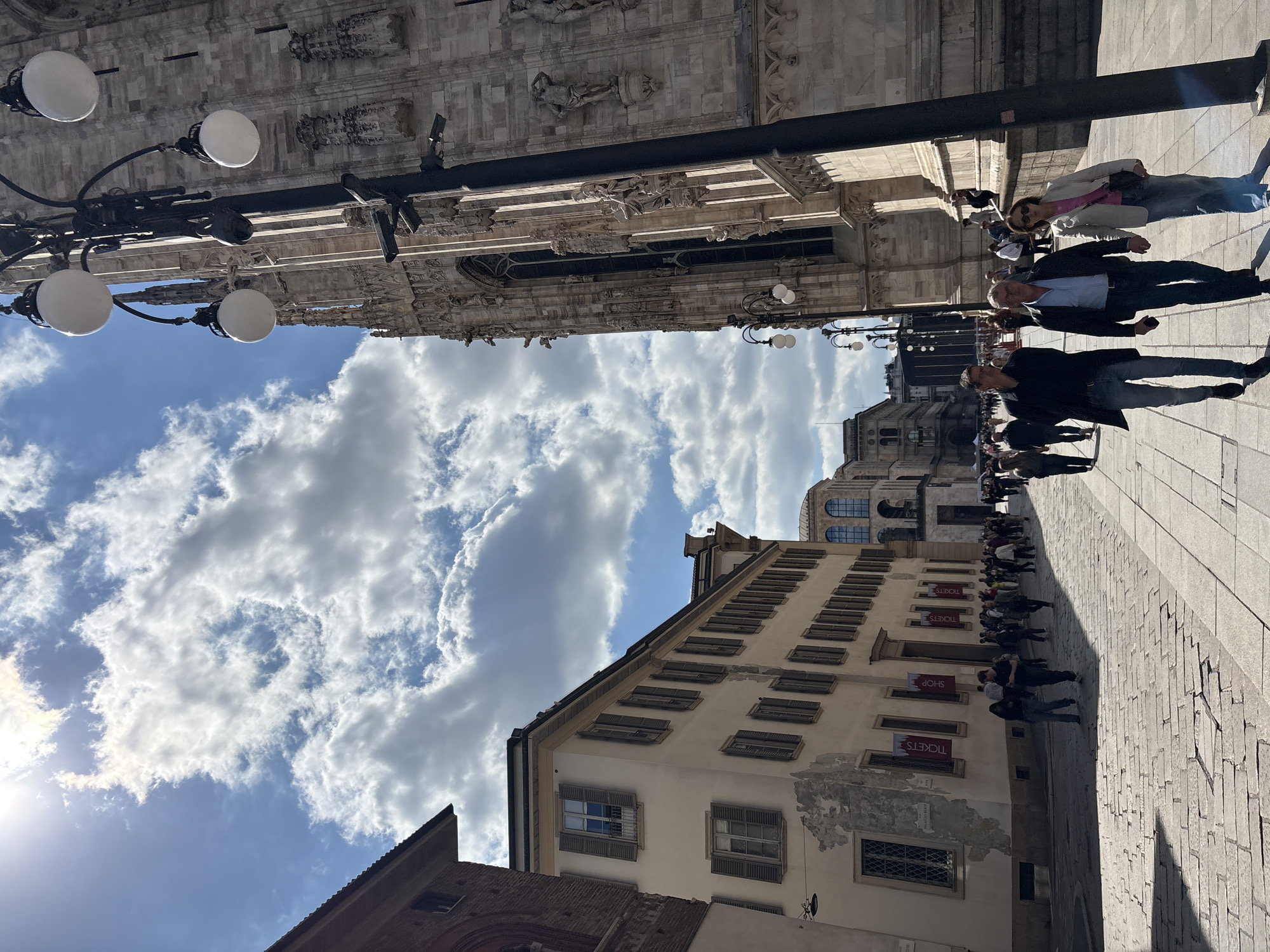 The image depicts a street scene in Milan, Italy, characterized by a mix of architectural styles and a partially cloudy sky. On the left, there is a building with a light-colored facade, multiple stories, and regularly spaced windows with shutters. Red banners with "TICKETS" are displayed. Along the right side of the image, people are walking on a cobblestone street. Architectural elements of the Duomo di Milano can be seen above, including ornate stonework and sculptures. Streetlights with spherical globes are visible in the foreground. The sky is blue with white clouds, and the sun is visible in the lower left corner.