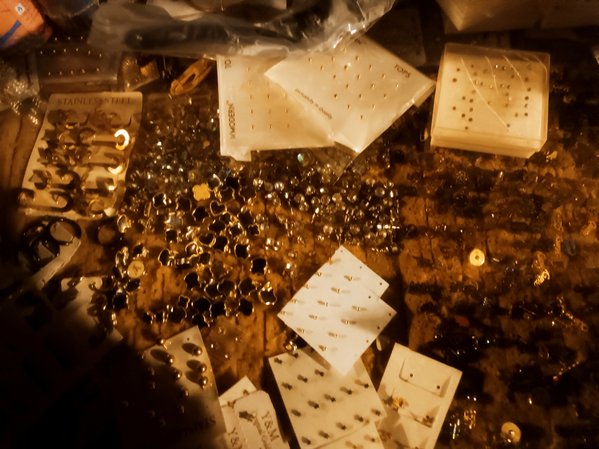 The image shows a close-up of various pieces of jewelry scattered on a surface, with cards holding earrings and other small adornments. The setting appears to be indoors with warm, yellowish lighting. A card reads "Stainless Steel" and another "Modern". The image appears to be taken in a shop that sells accessories. There are no people in the image, and no specific details about the environment suggest the location Bukuru, Nigeria. This identification cannot be verified.