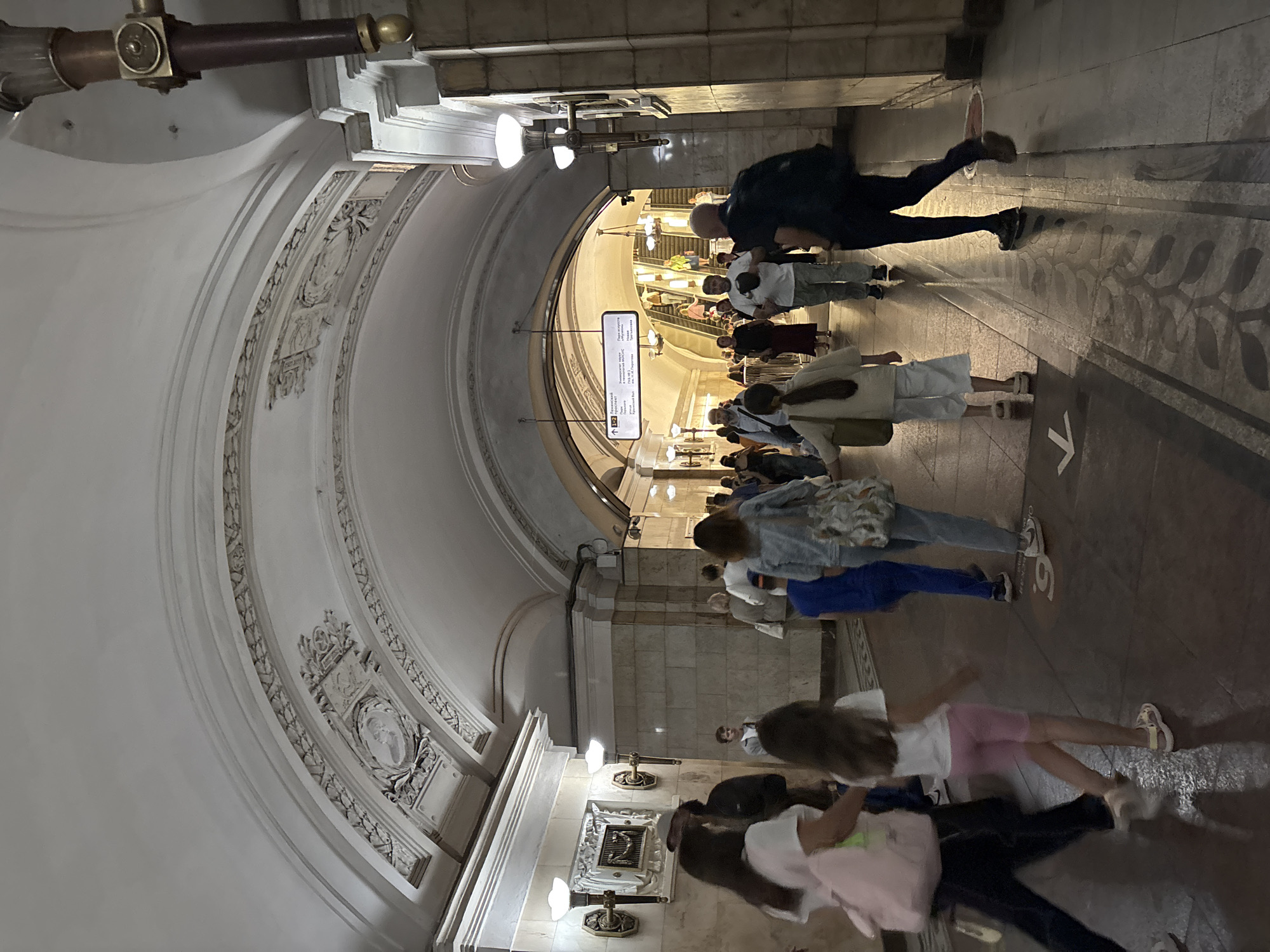 This image captures a scene within a grand, ornate metro station in Moscow, Russia, likely at a point where escalators are visible. The architecture features high, vaulted ceilings with intricate decorative moldings and relief sculptures, characteristic of many Moscow Metro stations. Several people are walking in different directions, some towards the escalators and others away from them.
In the foreground, on the left, a woman in dark pants and a white t-shirt is walking away from the camera, carrying a light pink backpack. Beside her, a young girl with long brown hair, wearing a white t-shirt and pink shorts, walks with her right leg extended forward. Further back, a child in a bright blue outfit is visible. To the right, a woman in a light blue denim jacket and a patterned skirt or dress walks towards the escalators.
The environment is well-lit by numerous wall-mounted lamps with ornate fixtures. The floor is paved with stone tiles, some of which have decorative patterns. A directional arrow pointing left is visible on the floor in the foreground. In the background, a large sign with text in Russian indicates directions and station names, and a set of escalators are ascending, with several people using them. The overall impression is one of a bustling, architecturally significant public transportation hub.