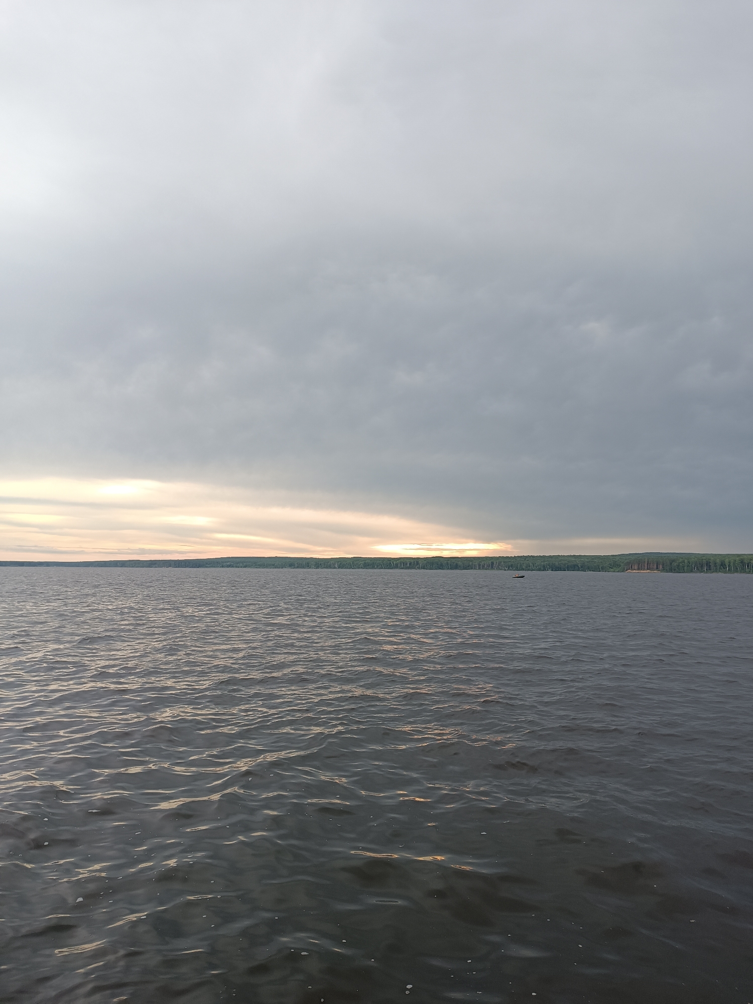 The image shows a wide, choppy body of water under a cloudy sky. A dense green forest lines the distant shore, with a break in the trees on the right side revealing a cleared area. A small boat with a single occupant is visible on the water in the middle distance. The sun is partially visible through the clouds on the horizon, casting a warm glow. The overall impression is of a serene, natural landscape. This scene is likely located on the Kama River in or near Chaikovsky, Russia, given the characteristic wide river and forested banks.