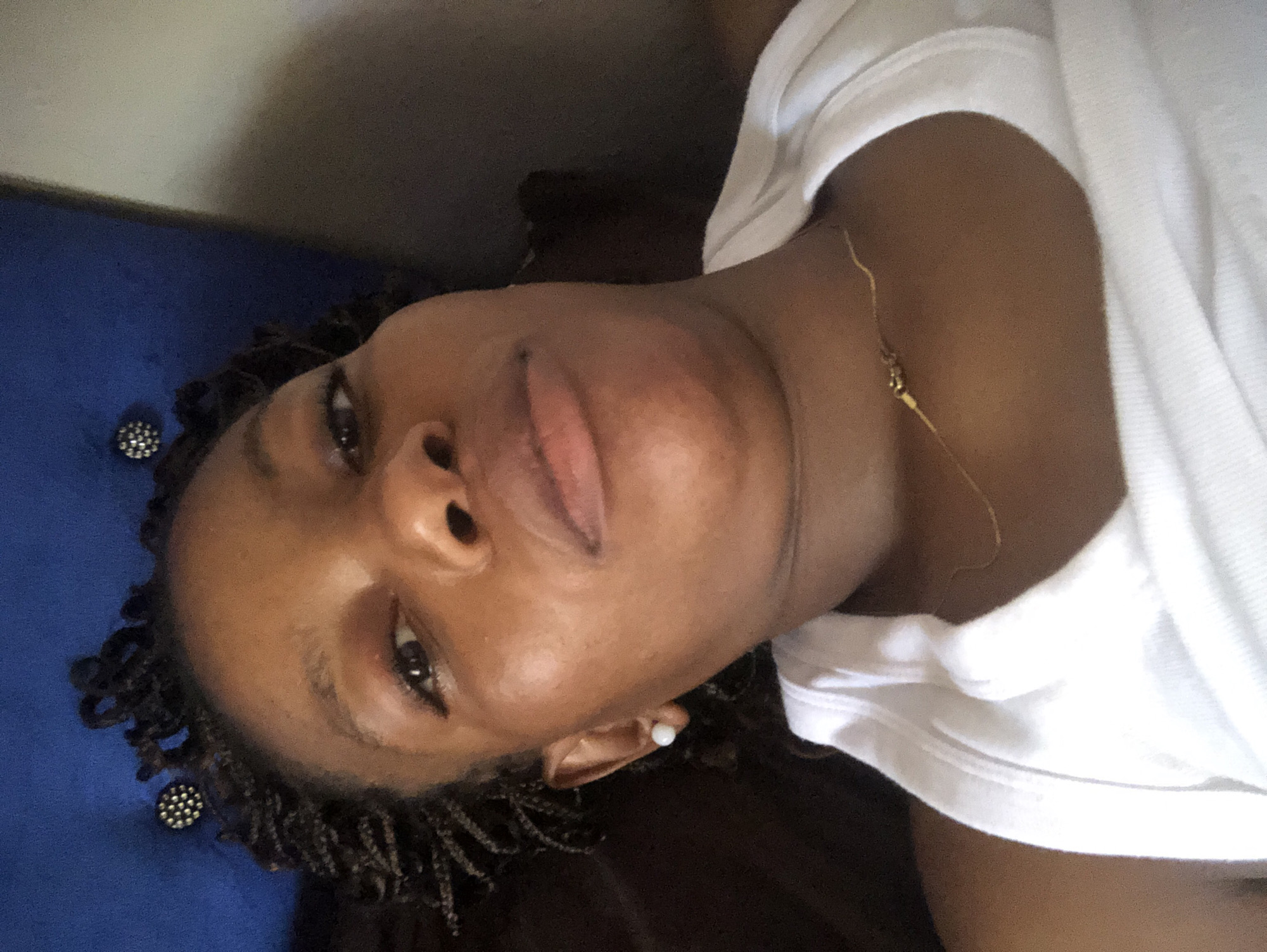 A person with dark skin is lying down, possibly on a blue tufted headboard, wearing a white t-shirt and a delicate gold chain. The person has styled hair and is wearing earrings. The location is presumed to be Kashere, Nigeria, though this cannot be verified from the image alone. There are no other people or discernible objects in the immediate view. The individual's activity and specific interactions are not evident. The environment is not further definable beyond the immediate surfaces the person is in contact with.
