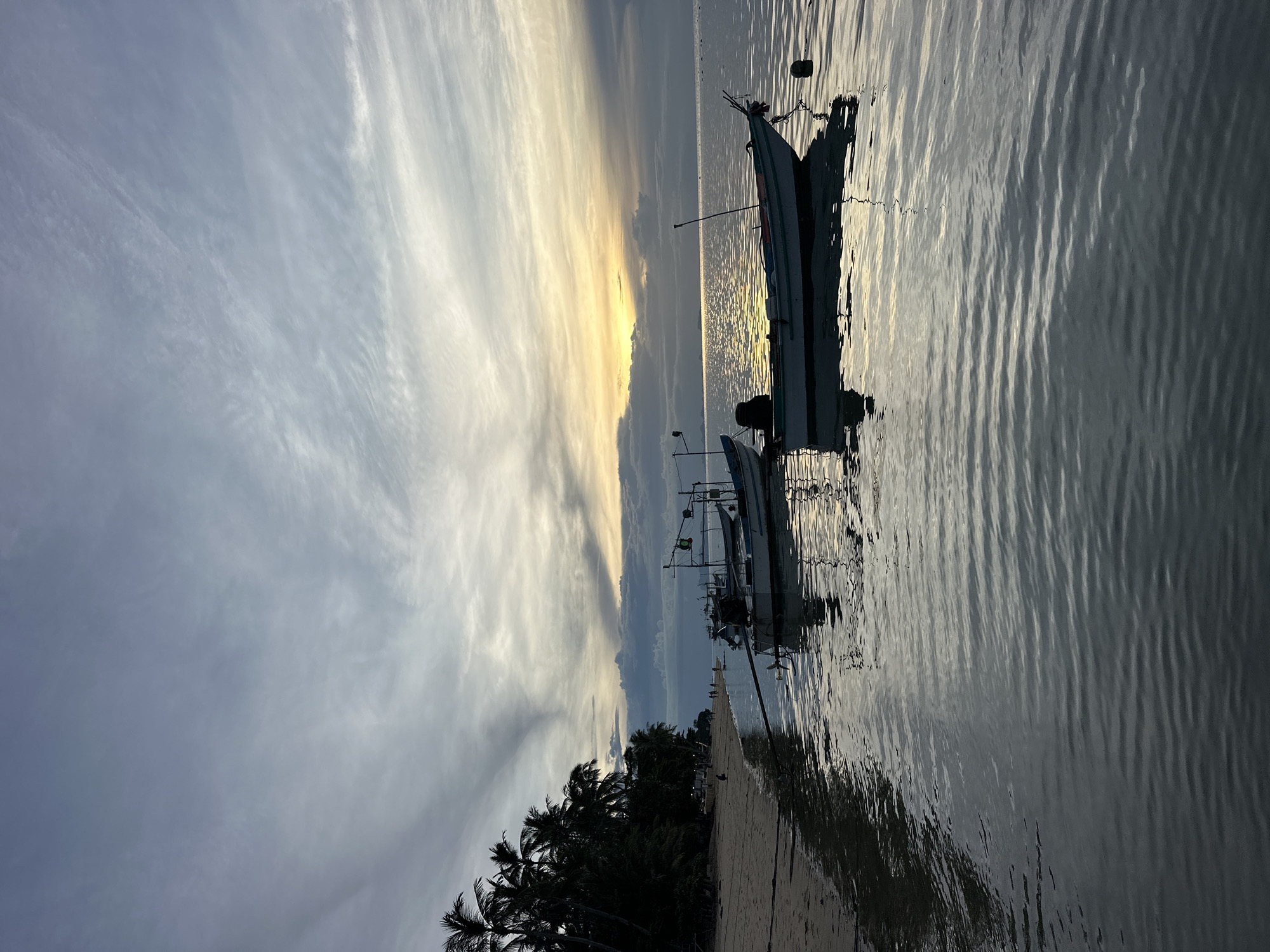 The image captures a serene beach scene at Baan Thong Po, Thailand, likely during sunrise or sunset. The sky is filled with a dramatic display of clouds, ranging from soft, wispy formations to darker, more solid masses, illuminated by the warm glow of the setting or rising sun on the horizon. A calm body of water reflects the sky's colors and the silhouettes of several small fishing boats anchored offshore.
On the left side of the image, a sandy beach is lined with a dense row of palm trees, their dark silhouettes stark against the lighter sky. The beach appears to be frequented by a few individuals, their presence indicated by small, dark shapes, suggesting a peaceful, uncrowded atmosphere. One person is visible sitting on the sand. Further down the beach, faint lights suggest structures or activity, but the overall impression is one of tranquility.
The water is calm, with gentle ripples creating patterns that mirror the sky and the boats. The boats themselves are simple, functional vessels, some with their motors visible. Their reflections are clear on the water's surface, adding depth and symmetry to the composition. The overall color palette is dominated by cool blues and grays in the sky and water, contrasted with the warm yellows and oranges of the horizon and the dark silhouettes of the trees and boats. The scene evokes a sense of peace and the natural beauty of a tropical coastline.
