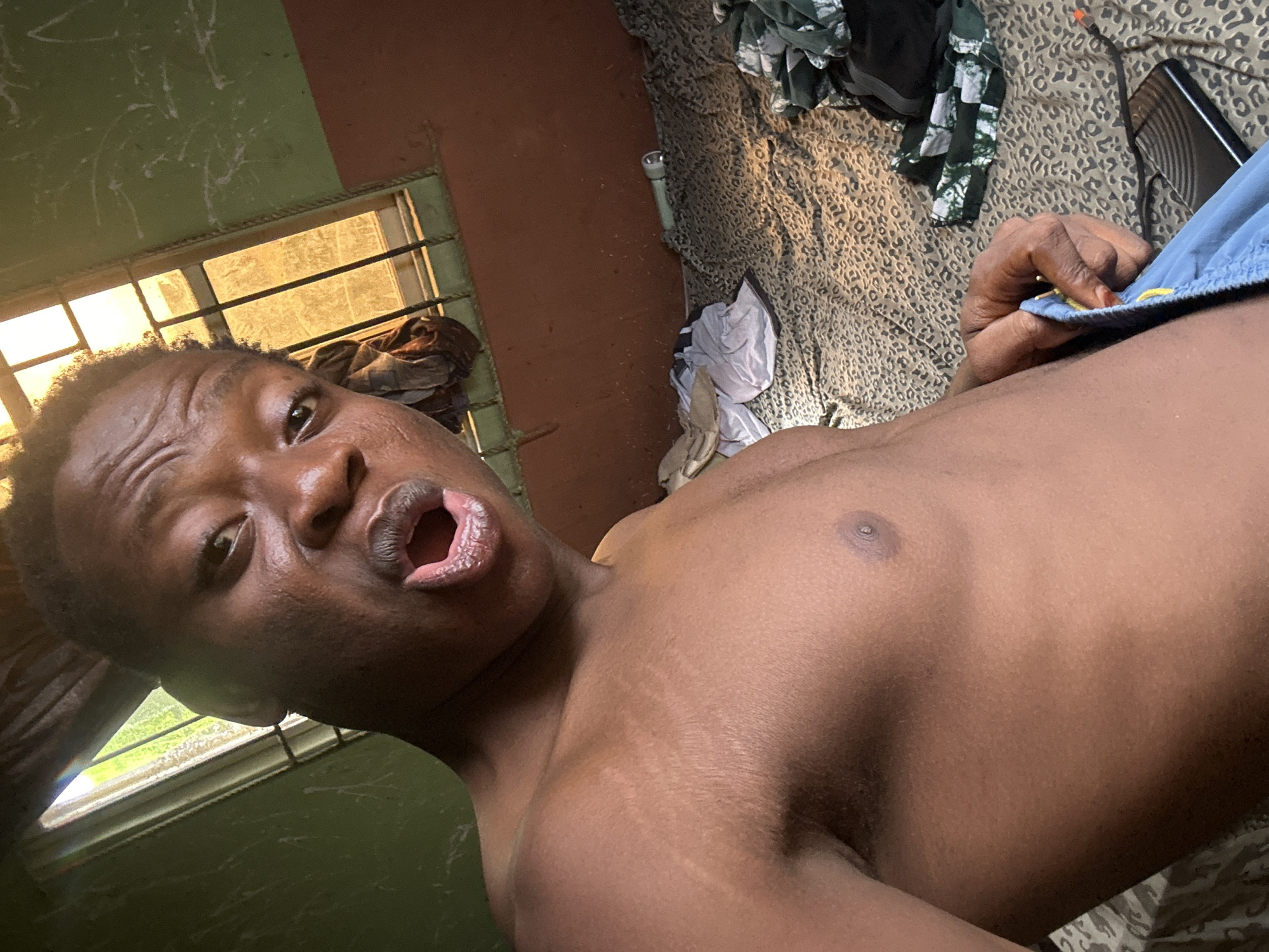 A close-up, low-angle shot captures a shirtless Black person, likely male, looking directly at the camera with an open mouth. Their short, dark, curly hair is visible, and their torso shows distinct stretch marks and appears somewhat shiny. Their right hand is seen holding the waistband of light blue shorts, which feature a yellow drawstring.
The setting appears to be indoors, possibly a bedroom. A window with dark metal security bars is visible on the left, letting in bright natural light, indicating it is daytime. The window illuminates a portion of the room's wall, which is painted in two sections: a lighter green upper half with white markings, and a darker reddish-brown lower half.
Behind the person, on what seems to be a bed or floor, there is a leopard-print fabric cover. Scattered on this fabric are various items, including a pile of green and black clothes, a white cylindrical flashlight, a black electronic device (possibly a phone) with an attached cable, and a small, light-colored animal-shaped object (possibly a plush toy or figurine). The overall impression is a candid, informal moment captured indoors. No visible text is present.
