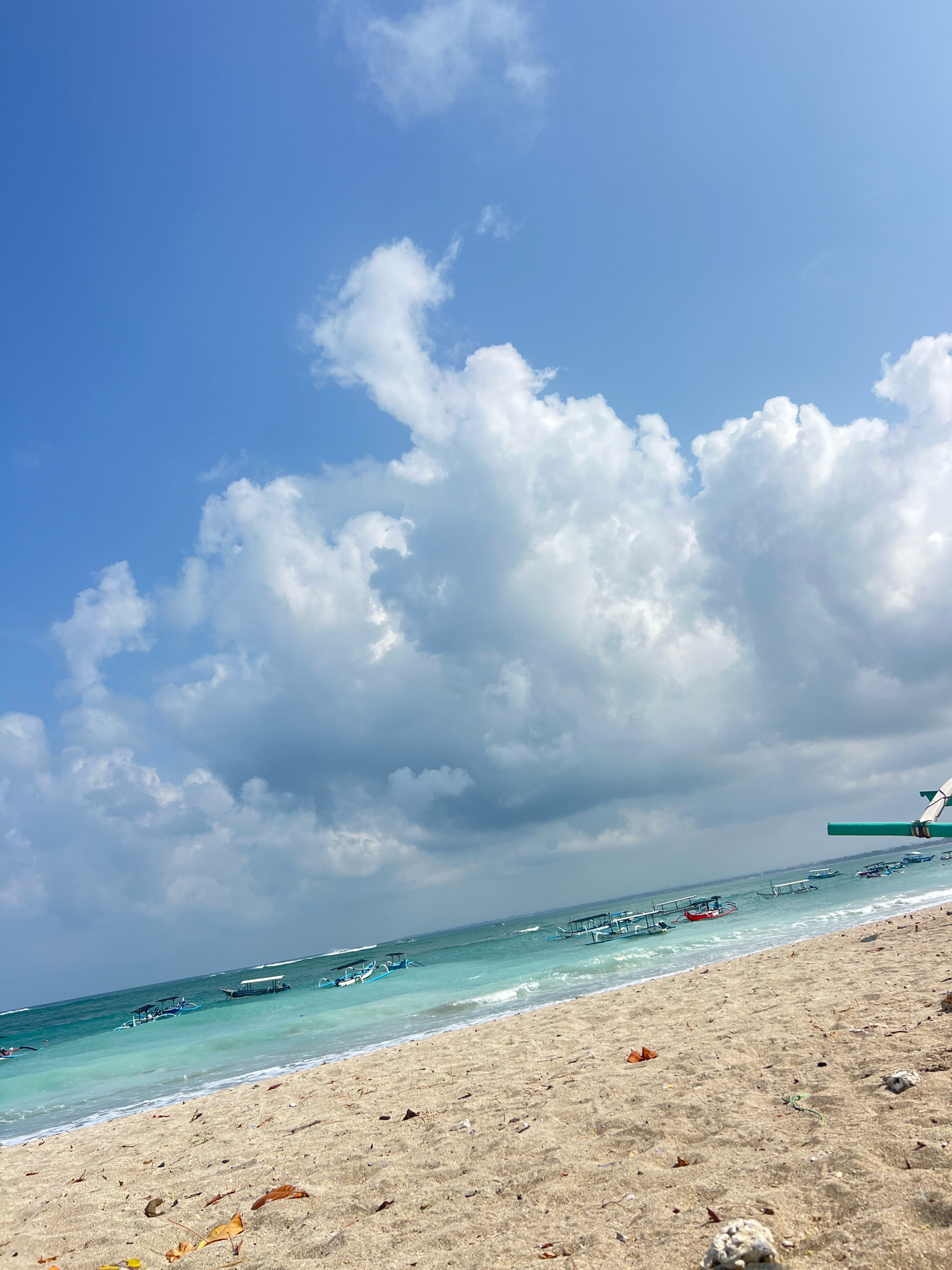 The image depicts a bright, sunny day at a beach in Kuta, Indonesia. The foreground is dominated by a sandy beach with scattered leaves and shells. The turquoise water of the ocean gently laps the shore, with small waves breaking. Several traditional Balinese fishing boats with outriggers are visible in the shallow water. In the background, a wide expanse of blue sky is dotted with fluffy white clouds, some of which appear darker and more ominous, suggesting a possibility of changing weather. The scene captures the essence of a tropical beach environment.