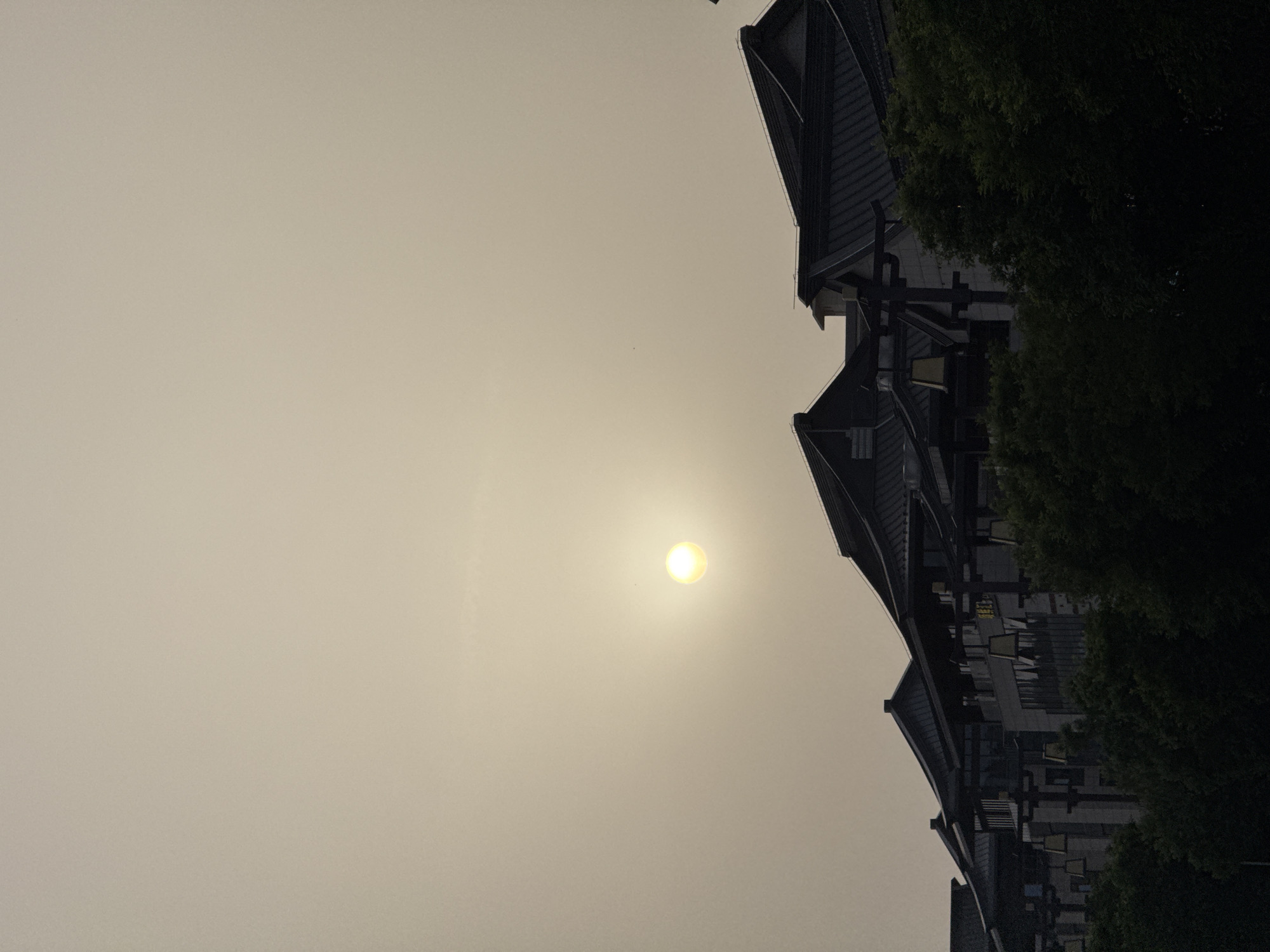 The image, rotated 90 degrees clockwise, shows a hazy sky with the sun visible as a bright orb. To the right, a building with multiple layers of roofs and some greenery is visible. The location is the city of Lintong District, China. There are no people visible in the image.