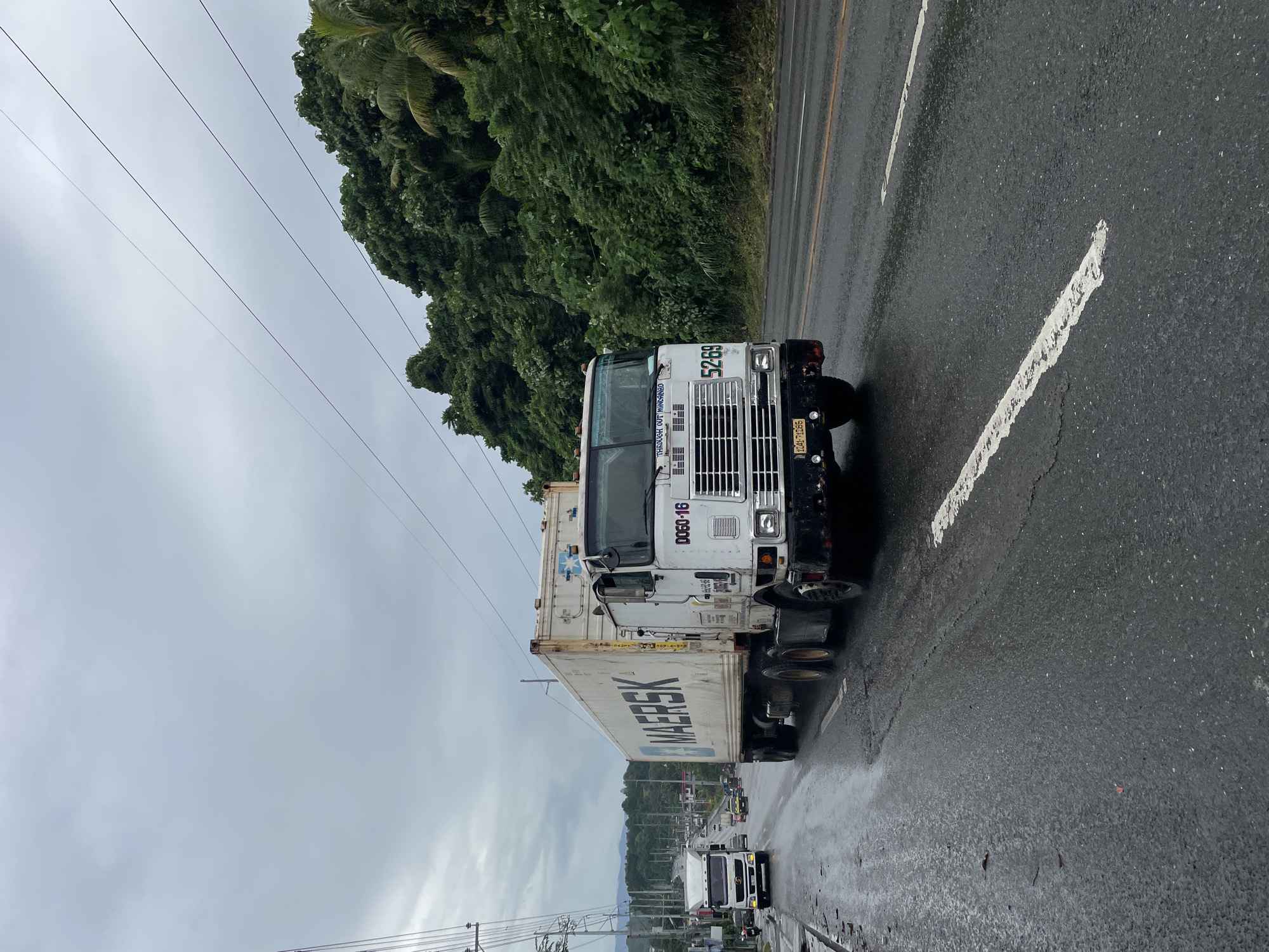 A white semi-trailer truck with "MAERSK" logo on the container is driving on a wet asphalt road with a cloudy sky. The truck is moving from left to right and is partially obscuring another white truck in the background. The road is lined with lush green trees on the right side, and there are power lines overhead. The overall scene appears to be a highway outside of a city. The license plate on the truck is partially visible and reads "1241-71285". The number "5269" is also visible on the front of the truck. The image was taken in Davao City, Philippines.