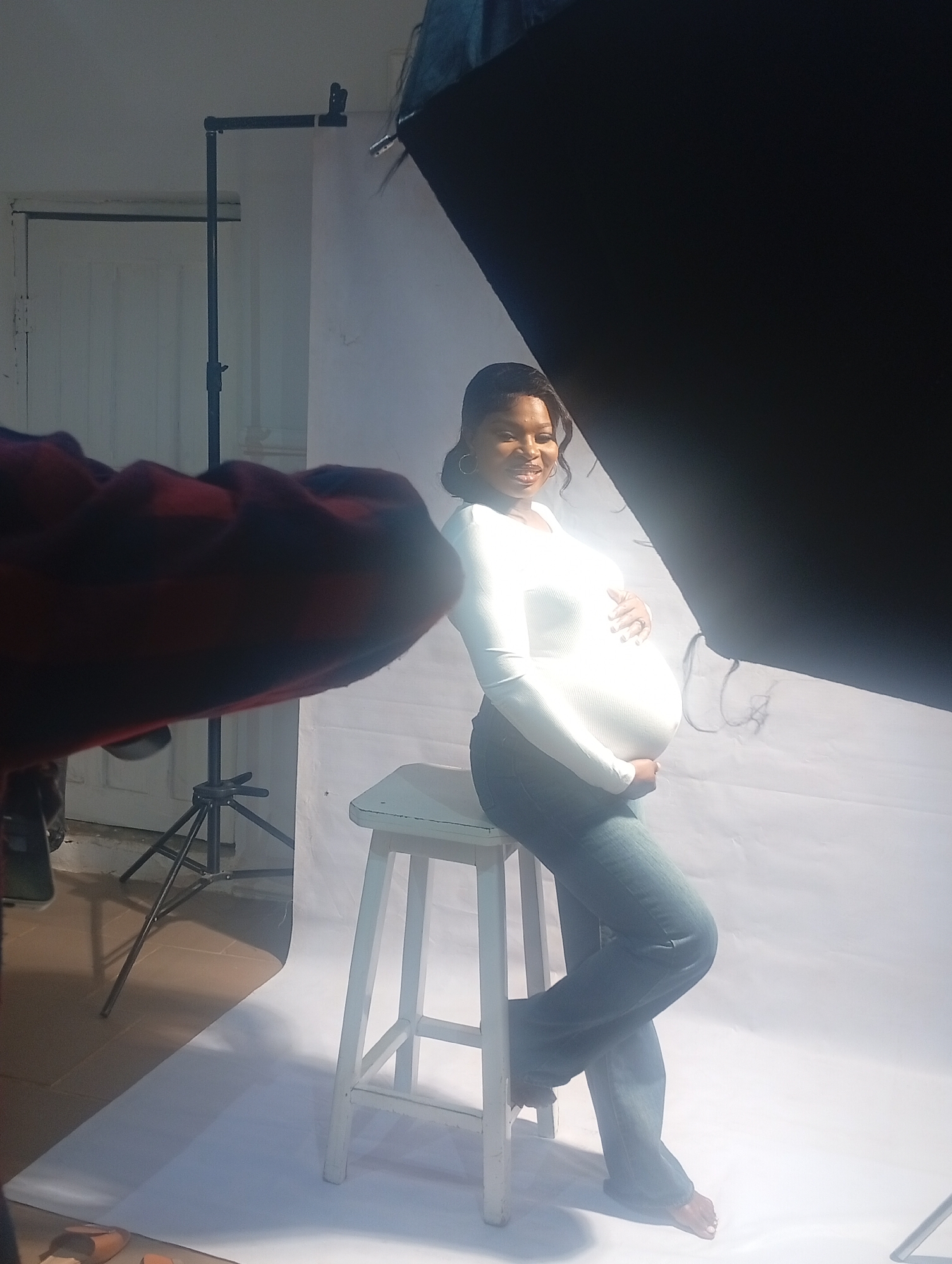 A pregnant woman is sitting on a white stool in a photography studio. She is wearing a white long-sleeved shirt and jeans, and she is holding her stomach. A large softbox is to the right of the woman, and another person, mostly out of frame, is to the left, possibly taking the photo. A light stand is visible in the background near a white door. The backdrop appears to be white. The photograph is taken in a studio setting in Jos, Nigeria.
