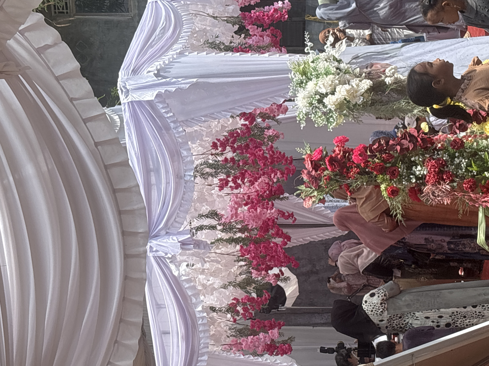 A wedding ceremony is taking place under a white tent adorned with pink and white flowers. Several individuals, mostly women wearing hijabs and modest attire, are gathered, some appearing to be in conversation or observing the event. Floral arrangements, including a prominent display of red and white flowers, are visible. In the foreground, two young girls are looking to the side. The scene appears to be an outdoor event in Lhokseumawe, Indonesia, with a textured grey wall and a window visible in the background. A camera on a tripod is also present, suggesting documentation of the occasion.
